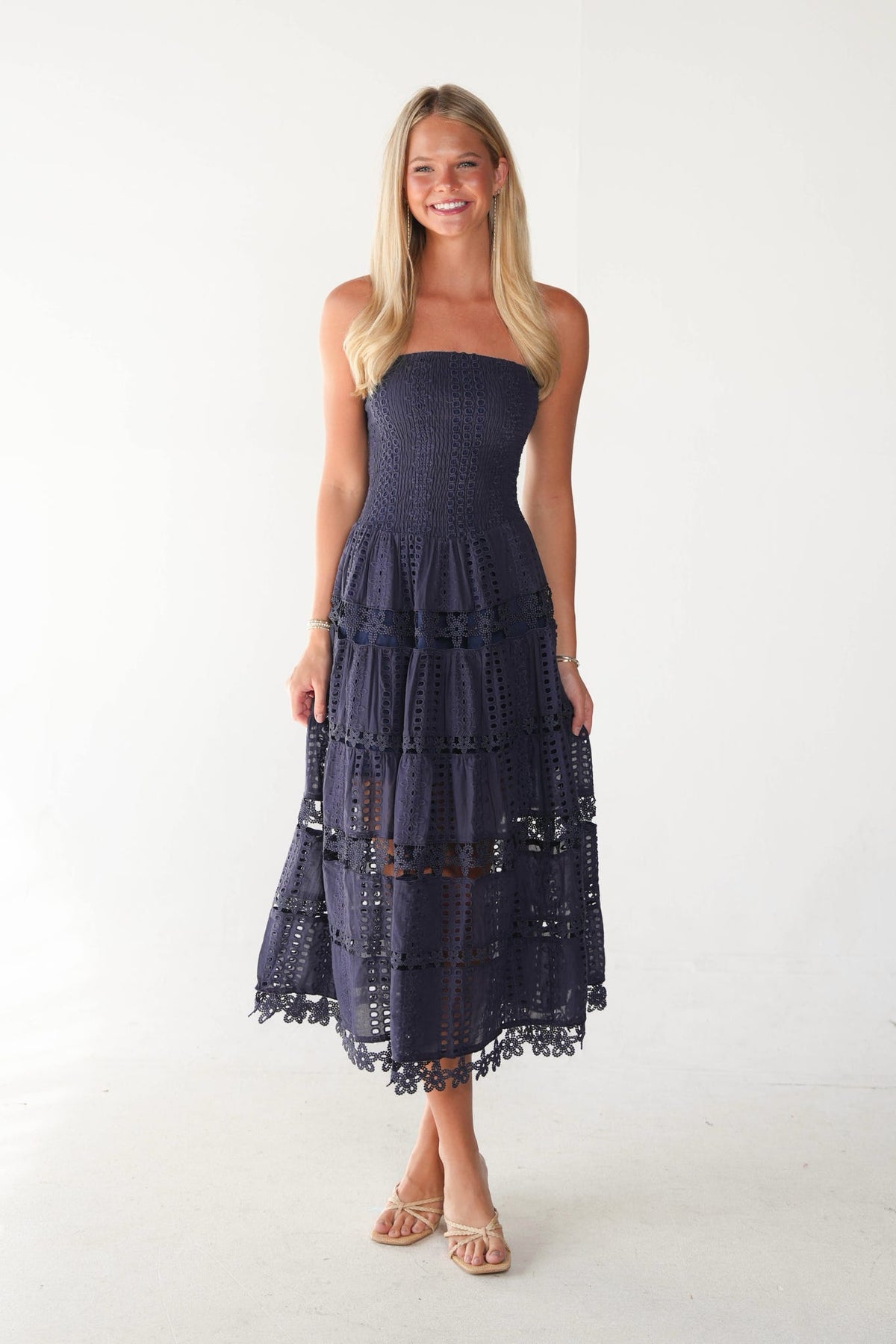 A woman modeling a strapless midi dress in a deep navy blue crochet or eyelet fabric, standing against a plain white studio background.