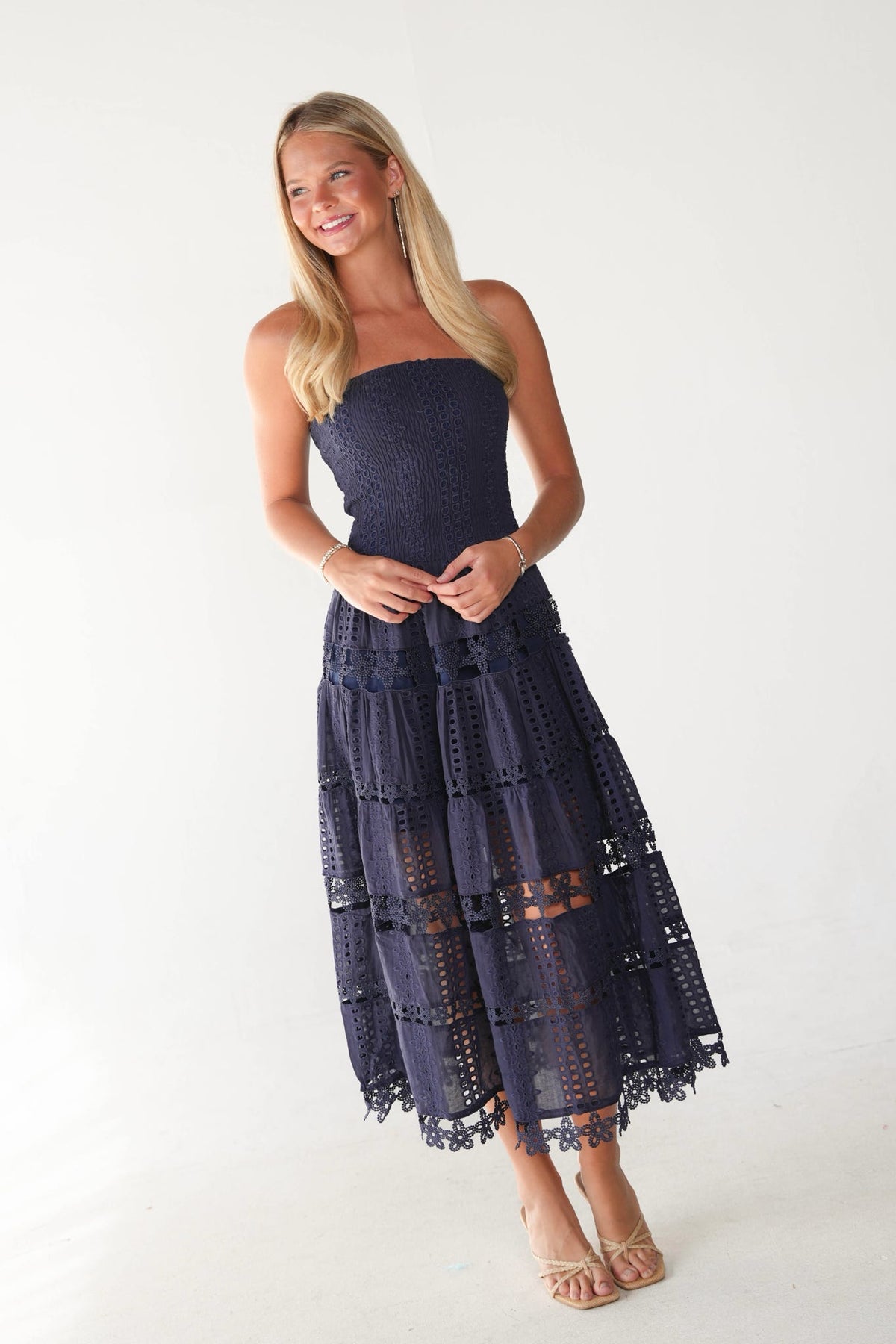 A woman modeling a strapless midi dress with a navy blue lace bodice and a tiered, eyelet lace skirt, photographed against a plain white studio background.