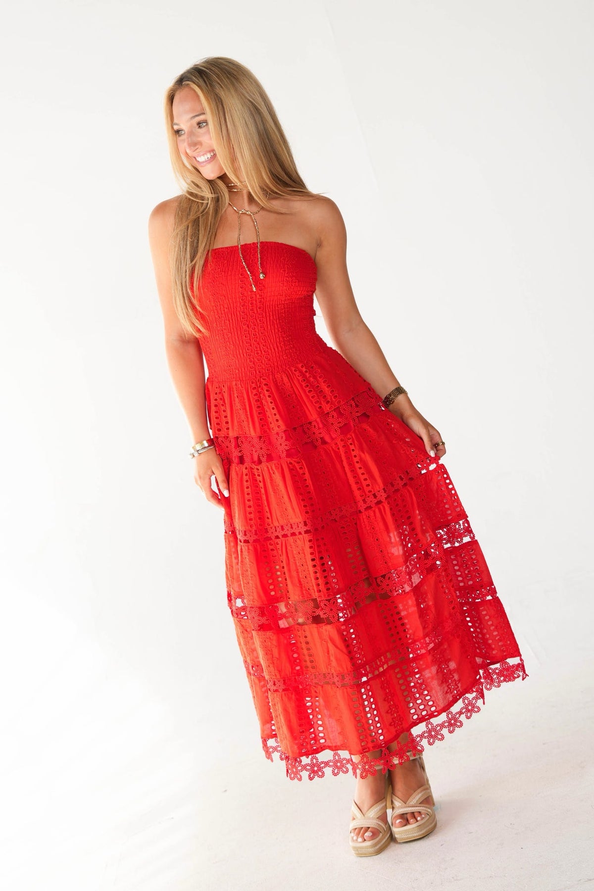 A woman modeling a bright red strapless midi dress with a tiered eyelet lace skirt, paired with beige strappy heels, against a plain white studio background.