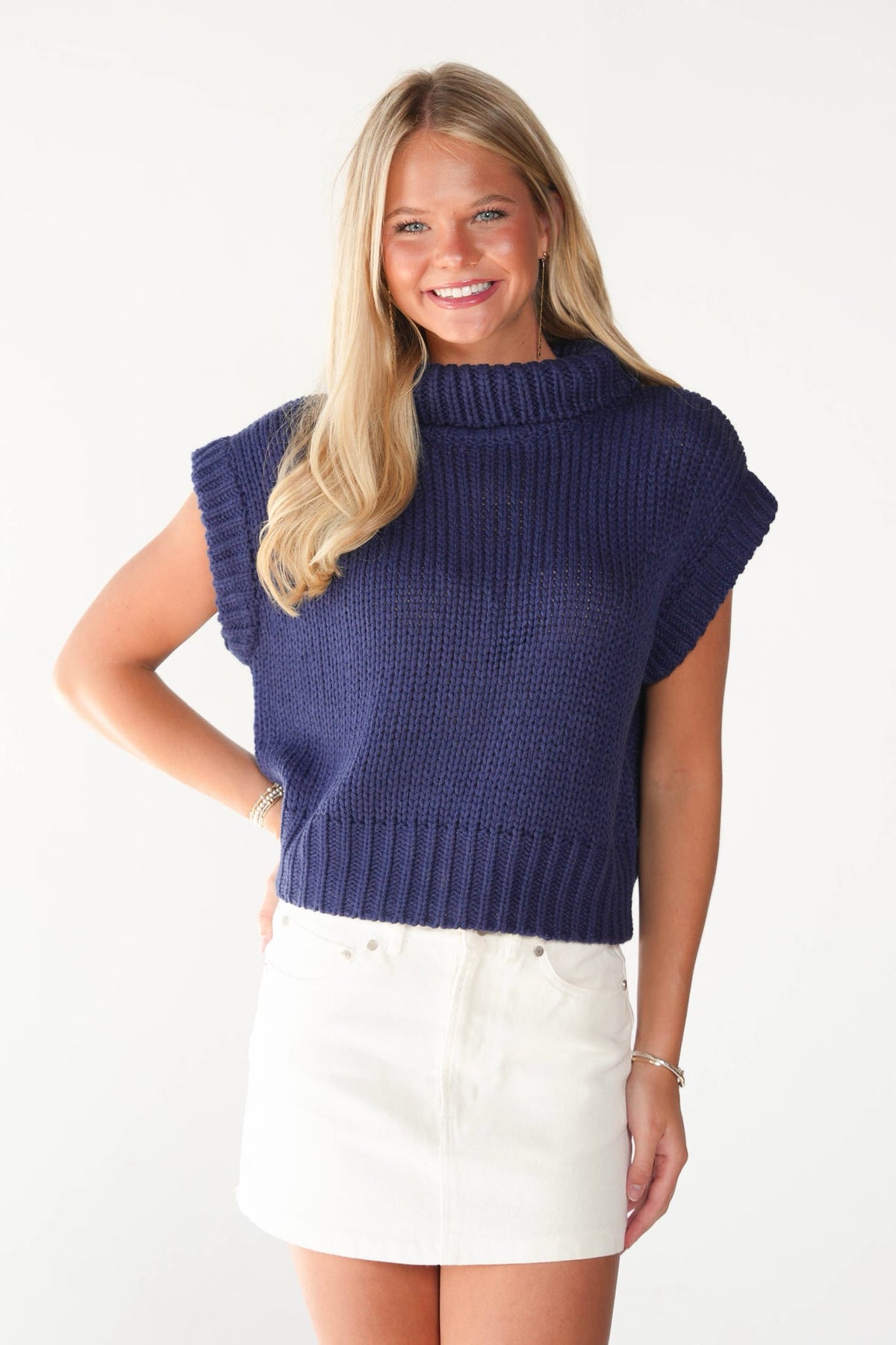 A smiling blonde woman wearing a navy blue ribbed knit mock-neck sweater vest with cap sleeves and a white mini skirt, standing against a plain white studio background.