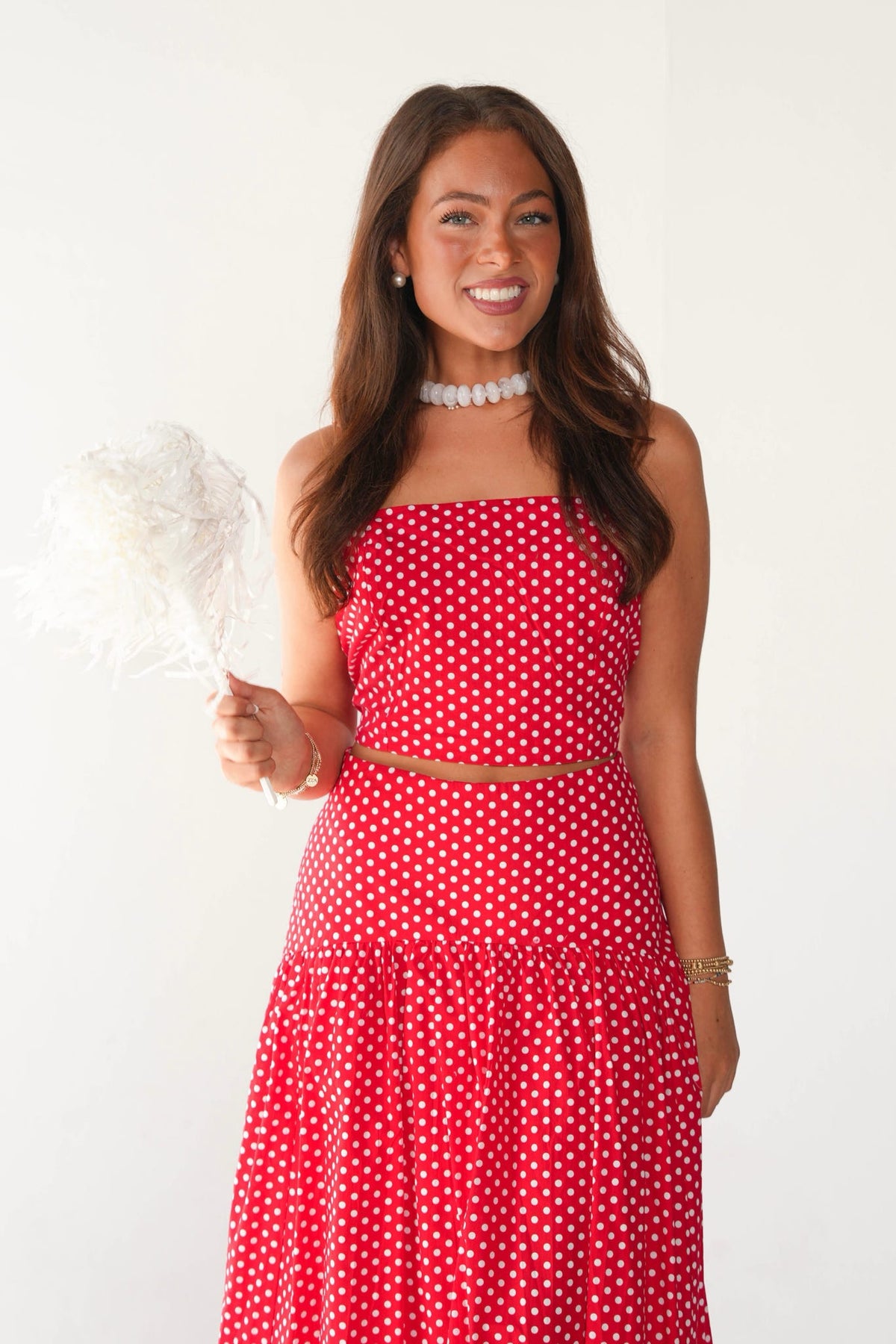 A woman modeling a red and white polka dot two-piece set consisting of a strapless crop top and a matching tiered skirt, holding a white feather fan against a plain white background.
