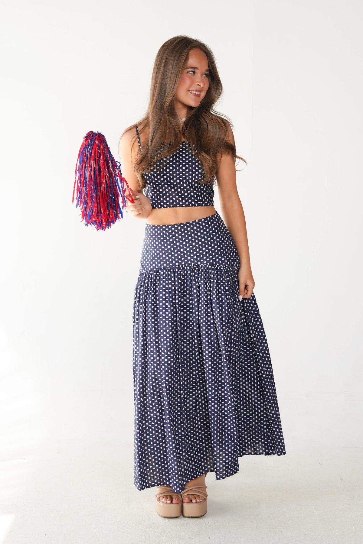 A woman modeling a navy blue and white polka dot two-piece set consisting of a halter crop top and a high-waisted flared skirt, holding a red and blue pom-pom against a white studio background.