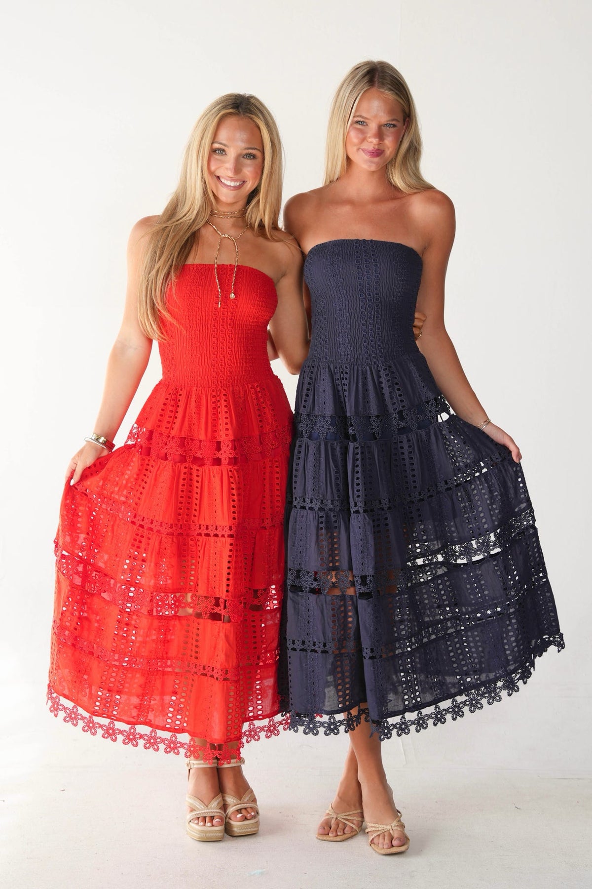 Two women modeling strapless eyelet maxi dresses against a plain white studio background. The woman on the left wears a bright red dress and holds out her skirt, while the woman on the right wears a dark navy blue dress and holds out her skirt.