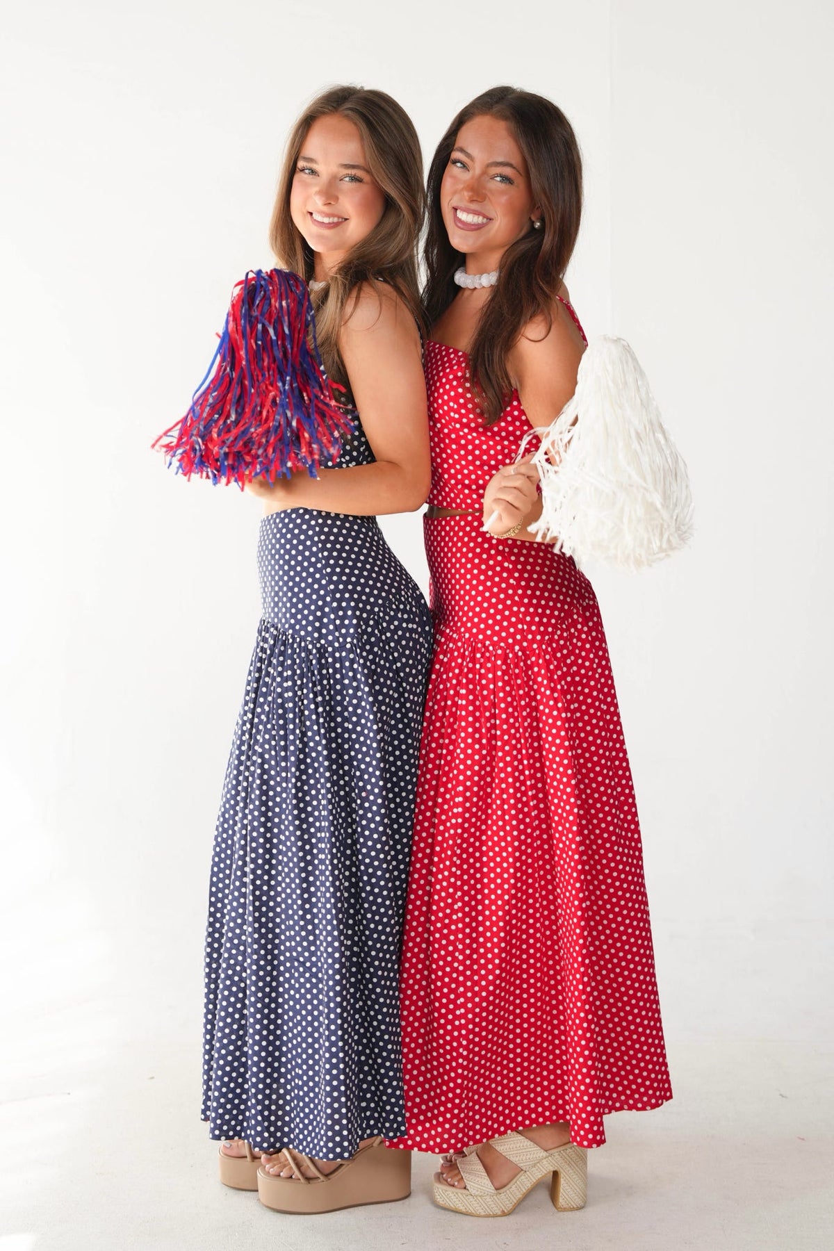 Two women modeling long white polka-dot maxi dresses in red and navy blue, standing against a plain white studio background. The woman on the left wears a navy dress with red and blue tassels, and the woman on the right wears a red dress with a white feathered accessory.