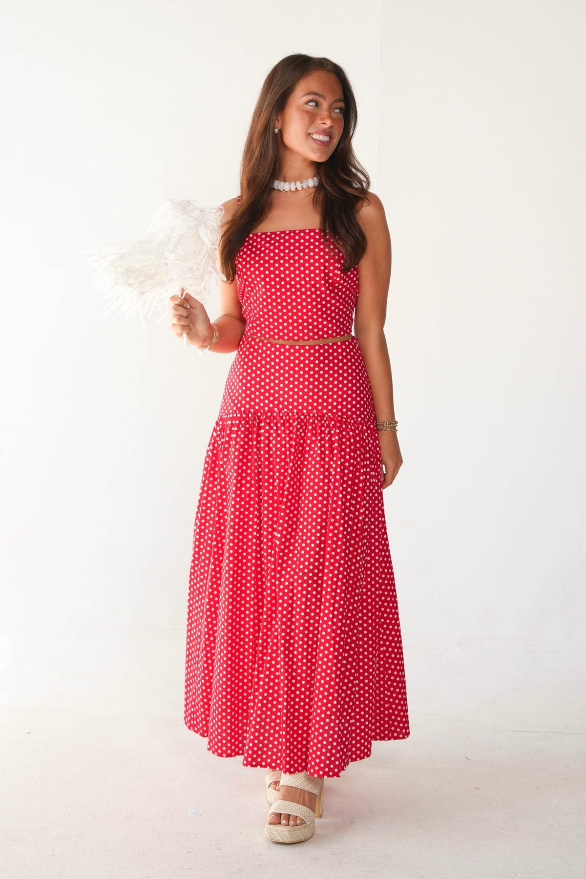 A woman modeling a red and white polka dot two-piece set consisting of a strapless top and a tiered maxi skirt, holding a white feather fan against a plain white background.