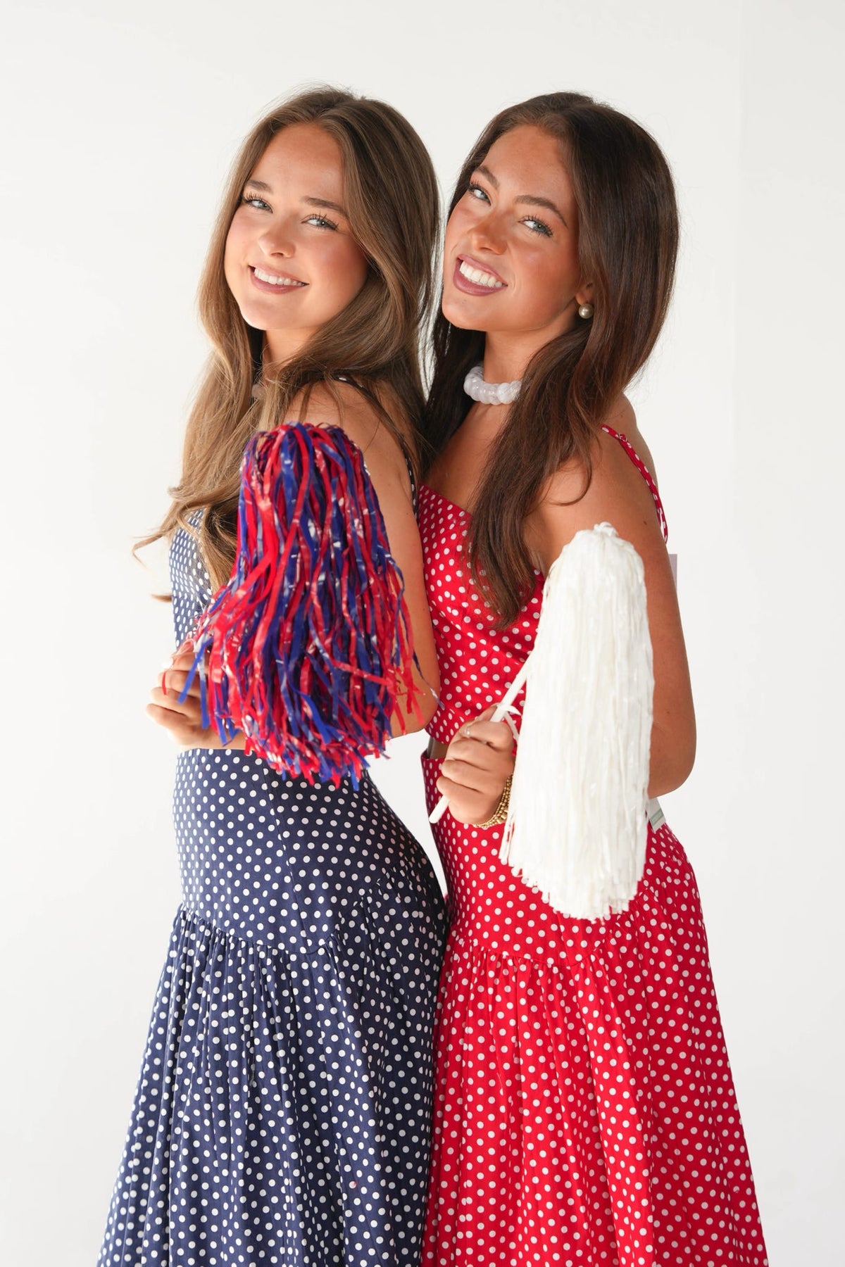 Two young women posing together against a plain white background, wearing red and navy blue polka dot dresses and holding patriotic red, white, and blue fringe accessories.
