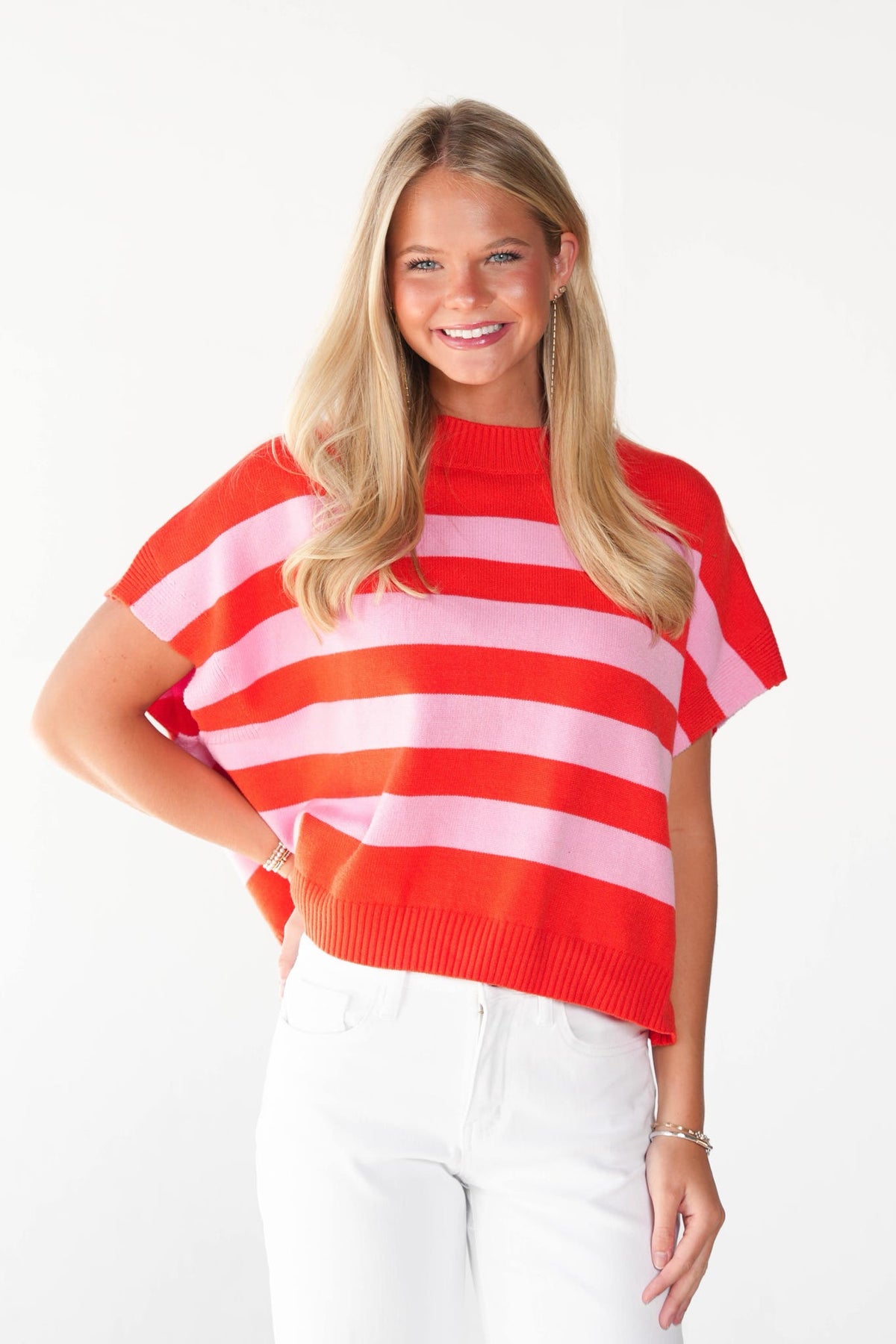 A smiling blonde woman wearing a short-sleeve knit sweater with bold horizontal red and pink stripes, paired with white pants, against a plain white studio background.