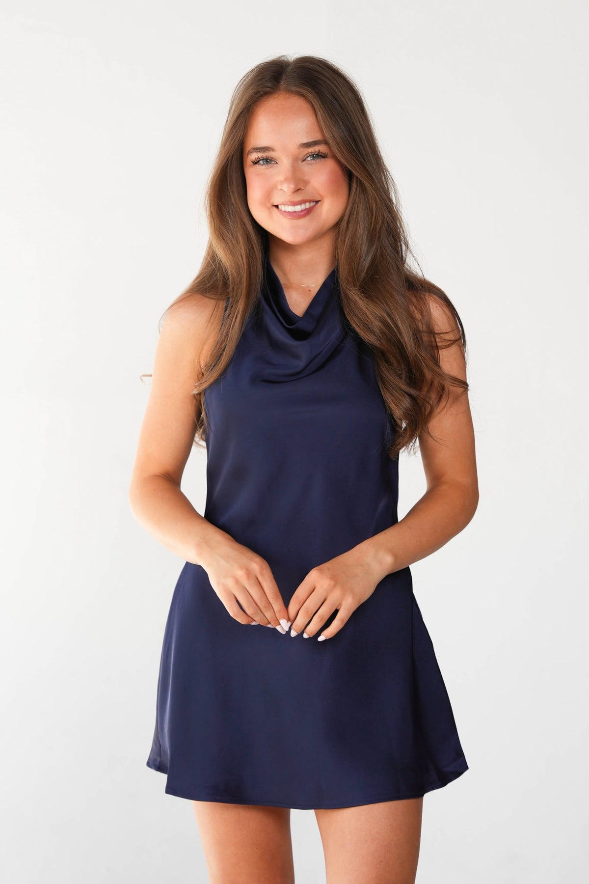 A young woman with long brown hair wearing a sleeveless navy blue cowl-neck mini dress, standing against a plain white studio background.