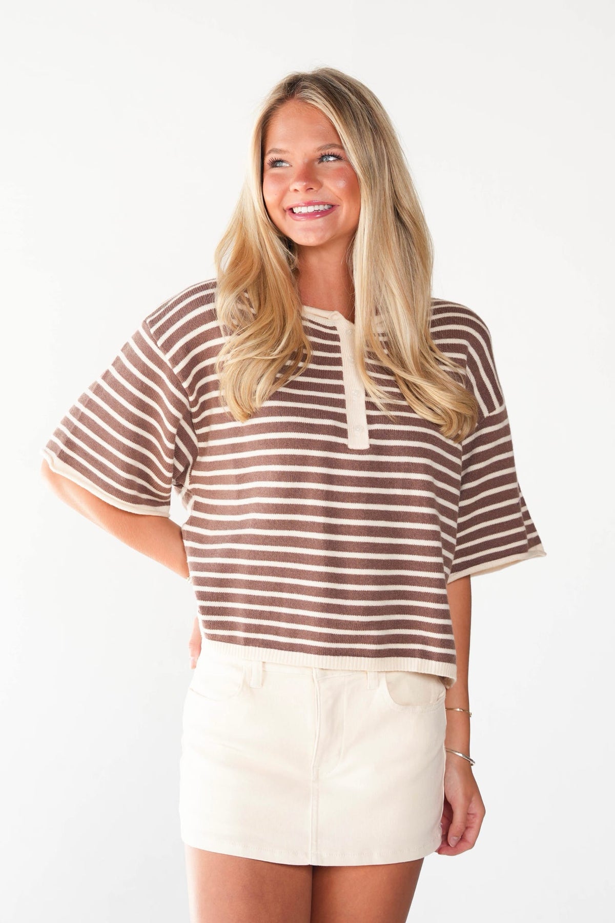 A smiling blonde woman wearing a brown and cream striped short-sleeve henley top with a tie-neck detail, paired with a cream mini skirt, against a plain white studio background.