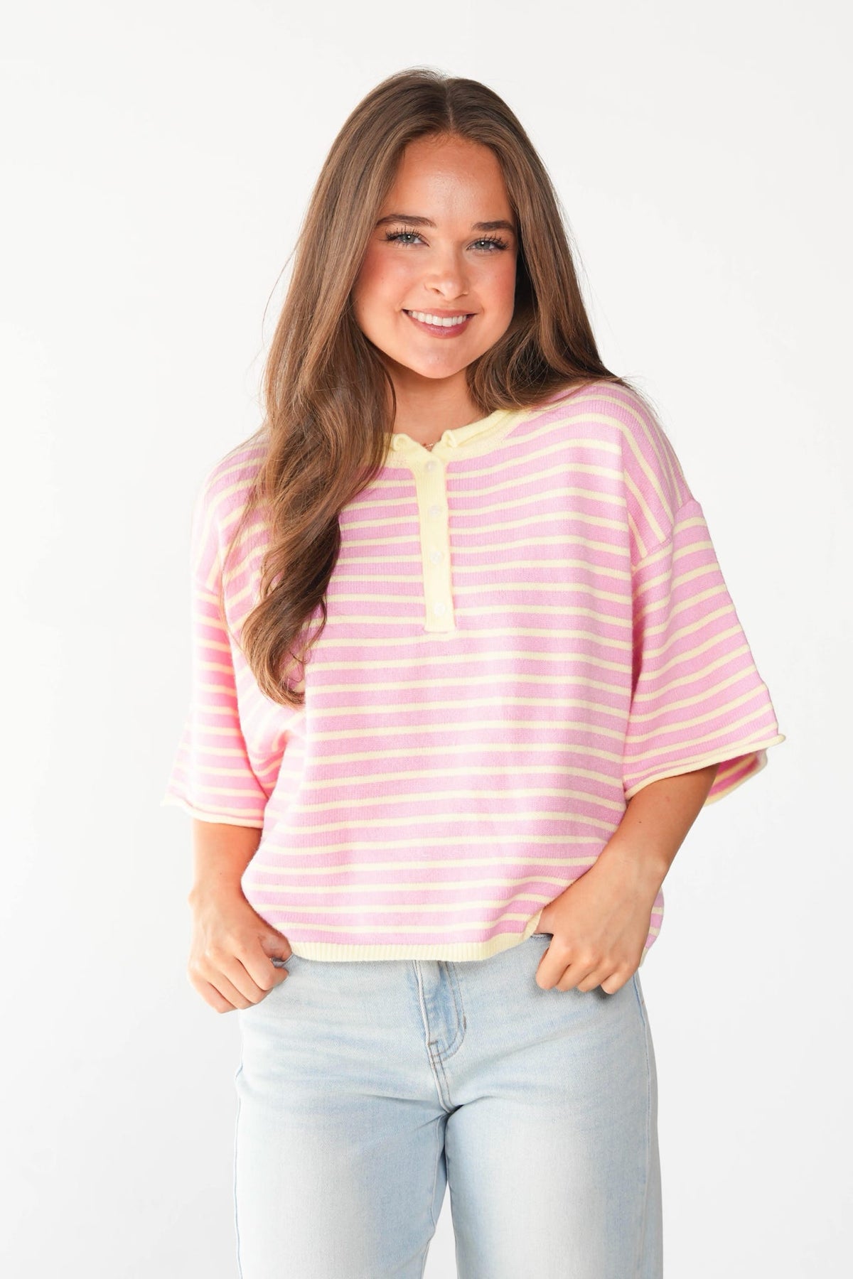 A young woman with long brown hair wearing a pink and white striped short-sleeve henley top with a yellow collar trim, paired with light blue jeans, against a plain white background.