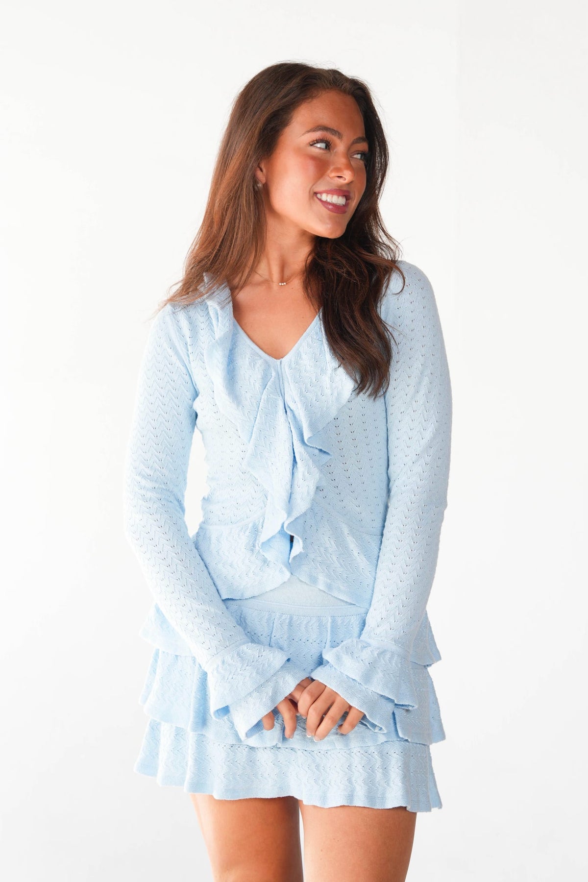 A woman modeling a light blue long-sleeve mini dress with a textured waffle pattern, ruffled tie neckline, and tiered ruffle hem against a plain white background.