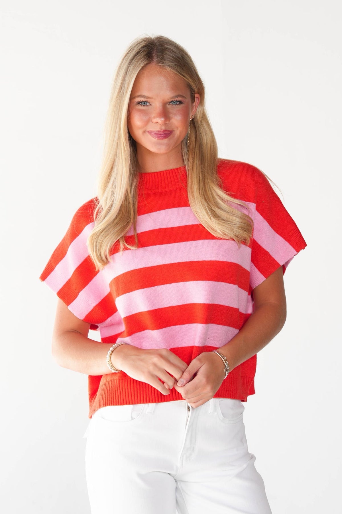 A blonde woman wearing a red and pink horizontally striped short-sleeve knit top with white pants, standing against a plain white background.