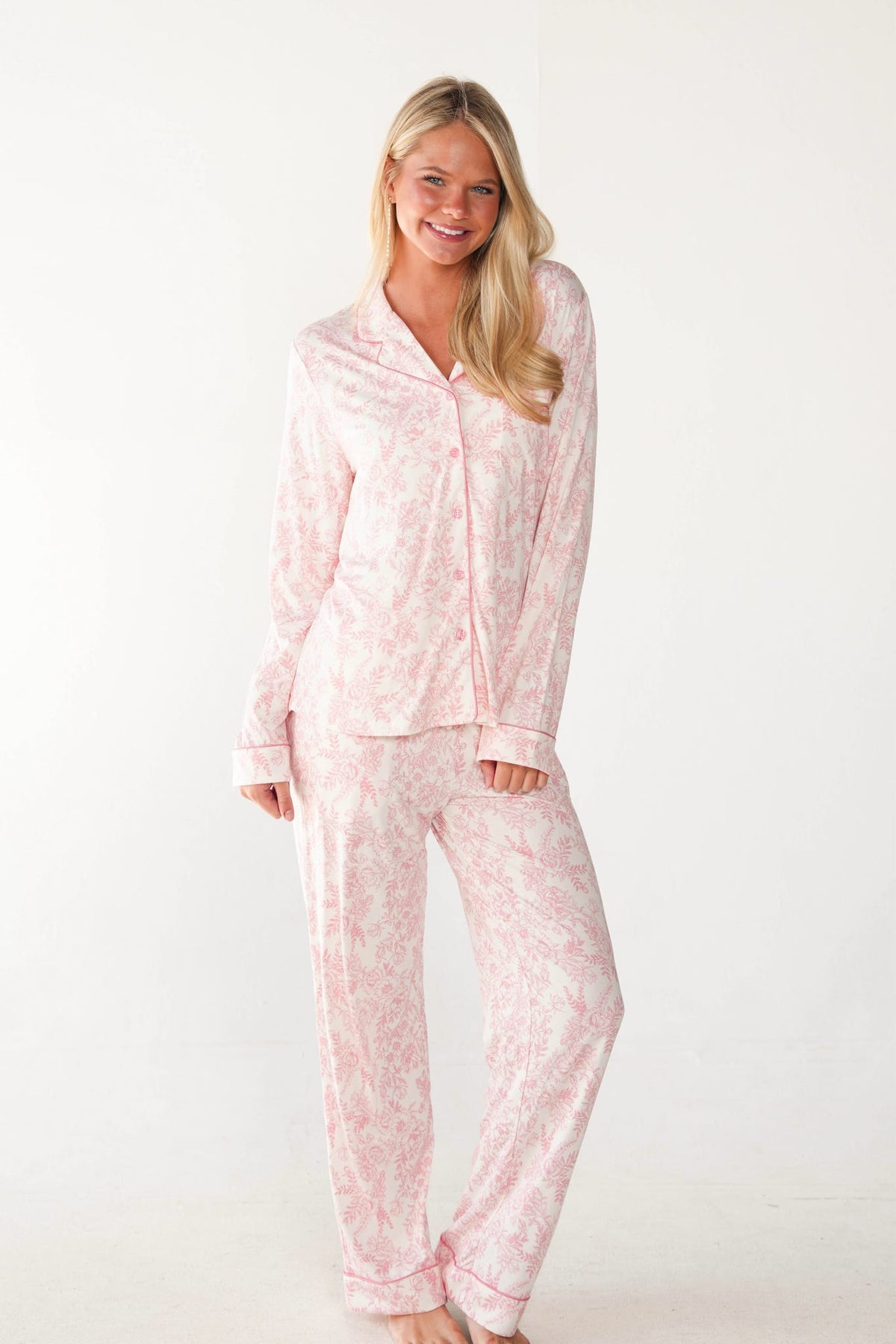 A woman modeling a light pink and white floral print pajama set, consisting of a long-sleeve button-up shirt and matching pants, against a plain white studio background.