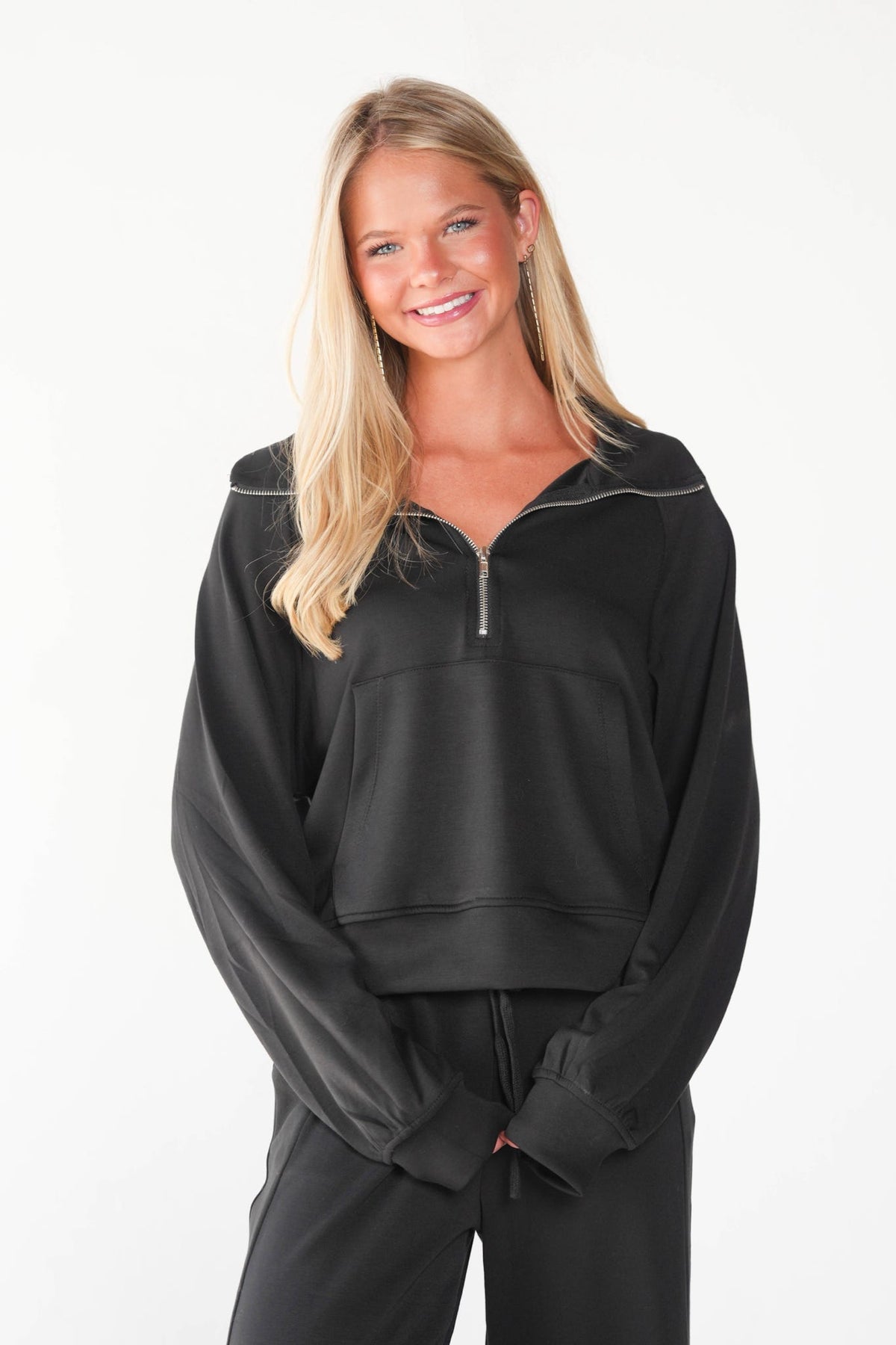 A smiling blonde woman wearing a black cropped half-zip sweatshirt with a half hood and black sweatpants, photographed against a clean white studio background.