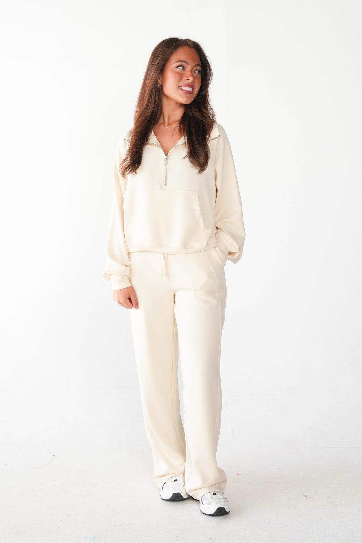 A woman modeling a cream-colored two-piece lounge set consisting of a half-zip pullover sweatshirt and matching wide-leg sweatpants, paired with white sneakers, against a plain white studio background.