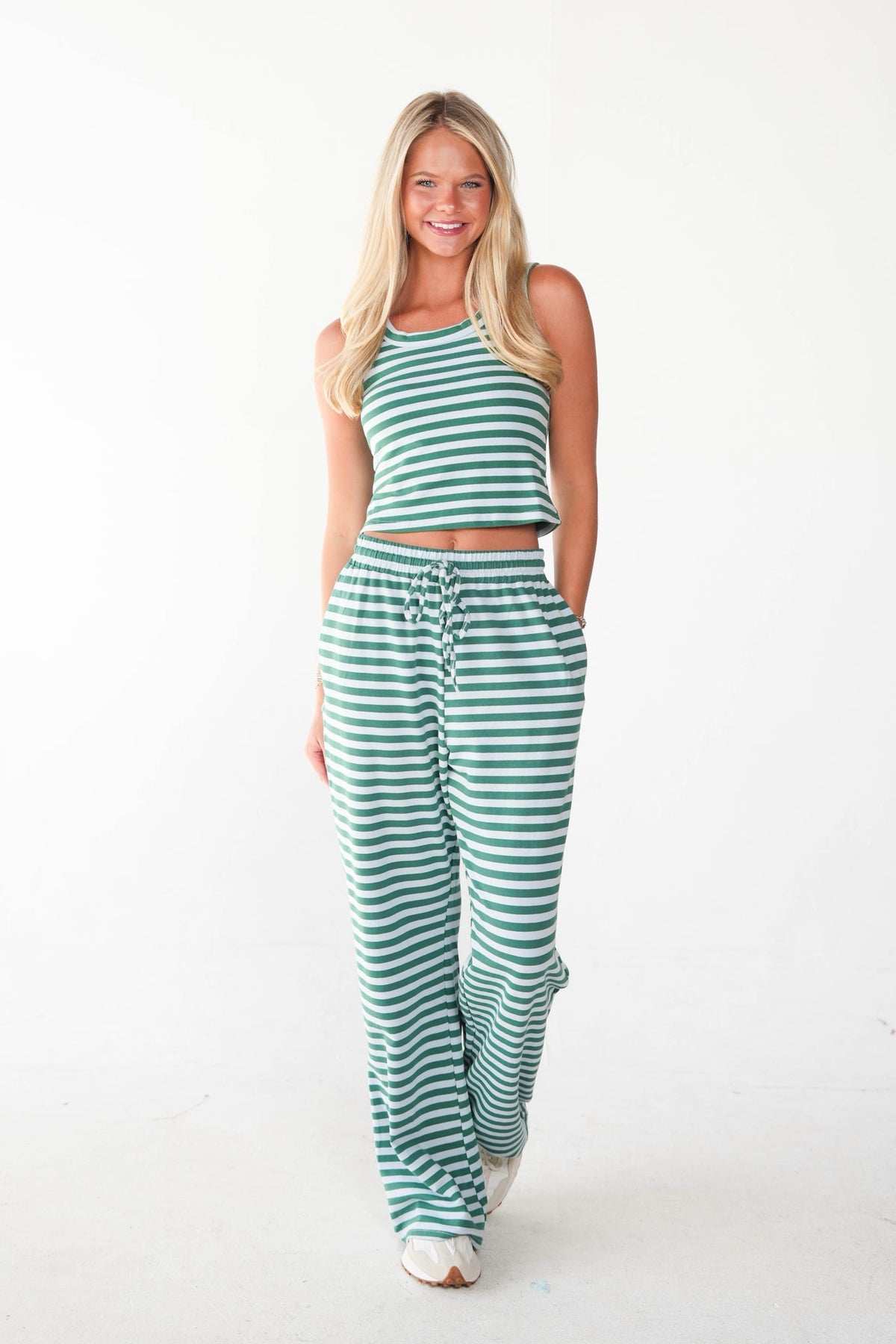 A woman modeling a matching two-piece loungewear set with green and white horizontal stripes, consisting of a halter crop top and drawstring sweatpants, against a plain white studio background.