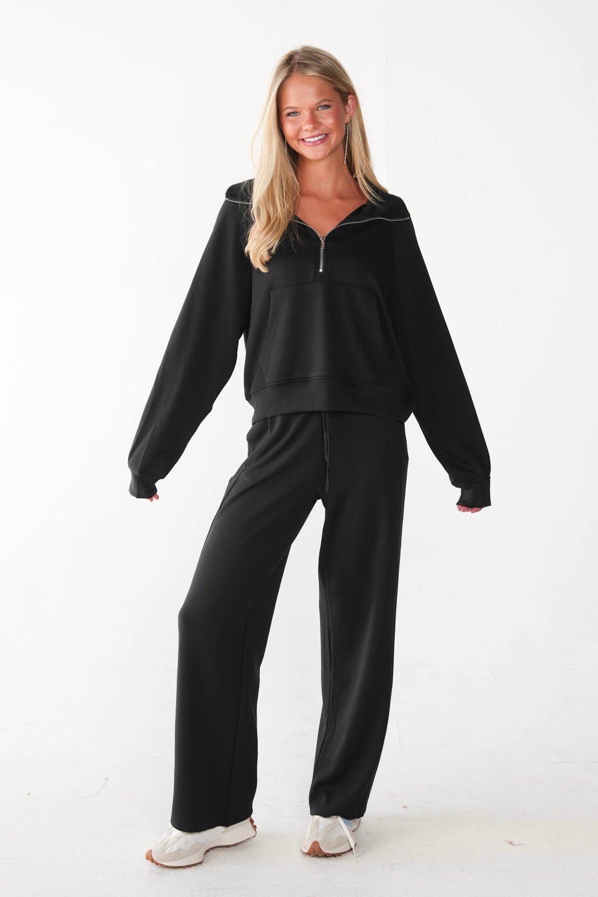 A woman modeling a black two-piece lounge set consisting of a half-zip sweatshirt and matching wide-leg sweatpants, paired with white sneakers, against a plain white studio background.