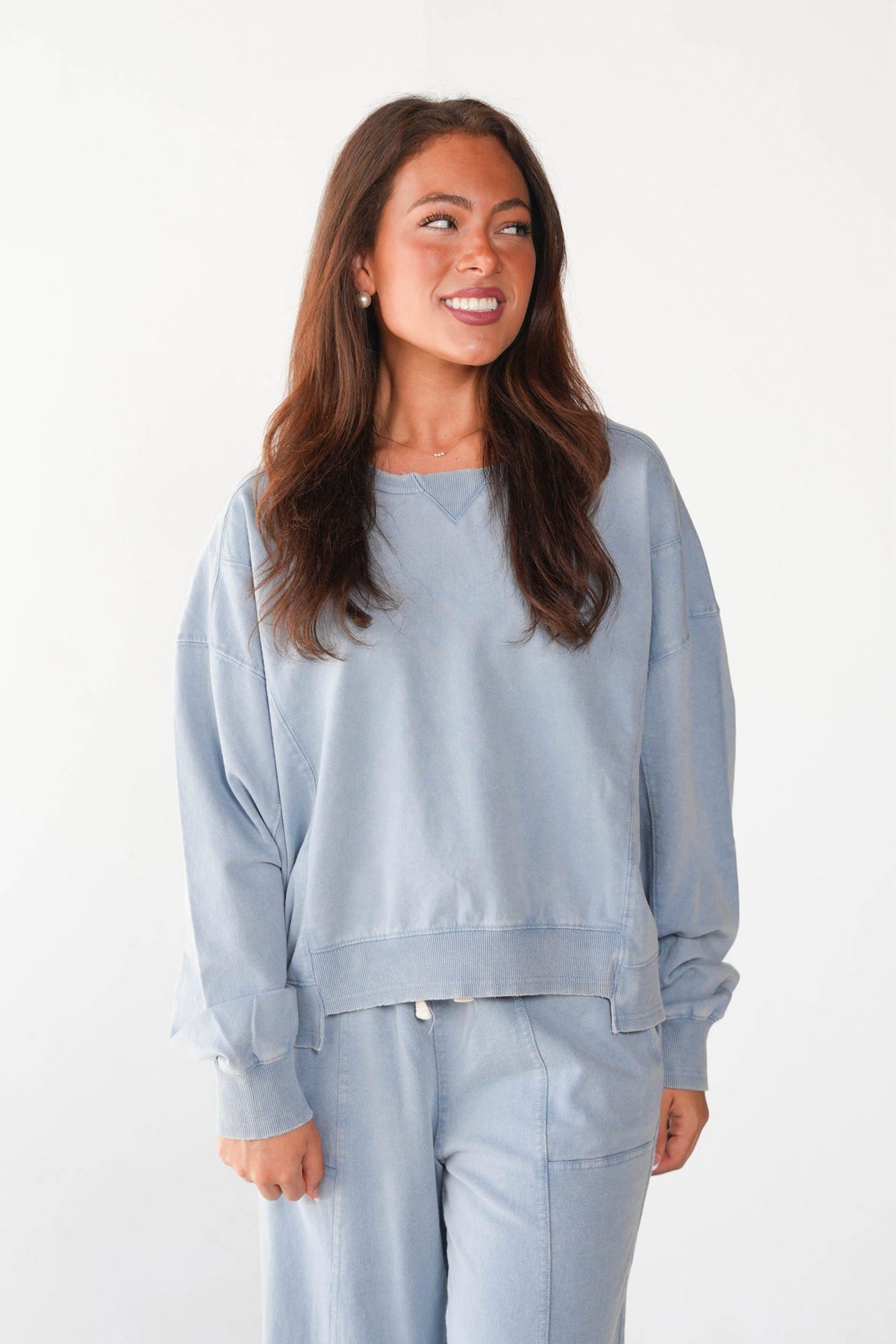 A woman wearing a light blue sweatshirt and matching sweatpants, standing against a plain white studio background.