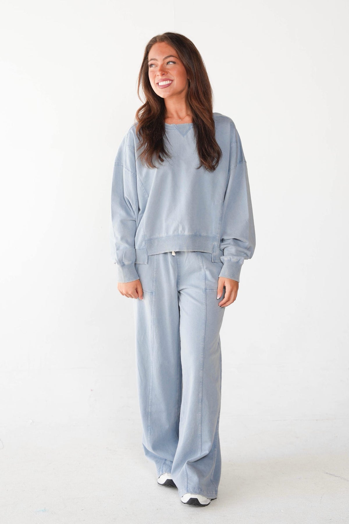 A woman modeling a matching light blue lounge set consisting of a crewneck sweatshirt and wide-leg pants, photographed against a seamless white studio background.