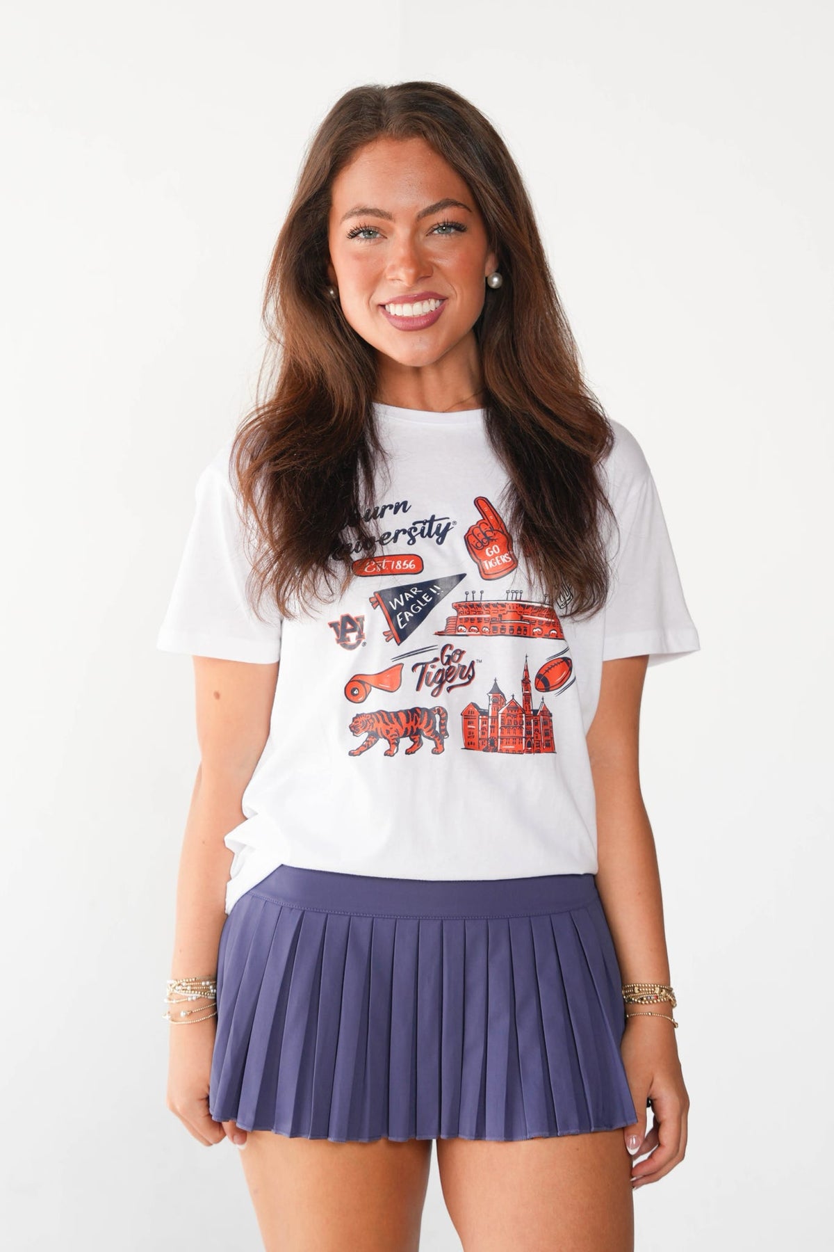 A smiling woman with long brown hair stands against a light gray studio background wearing a white graphic T-shirt and a pleated purple tennis skirt. The T-shirt features red and navy collegiate-style graphics and text.