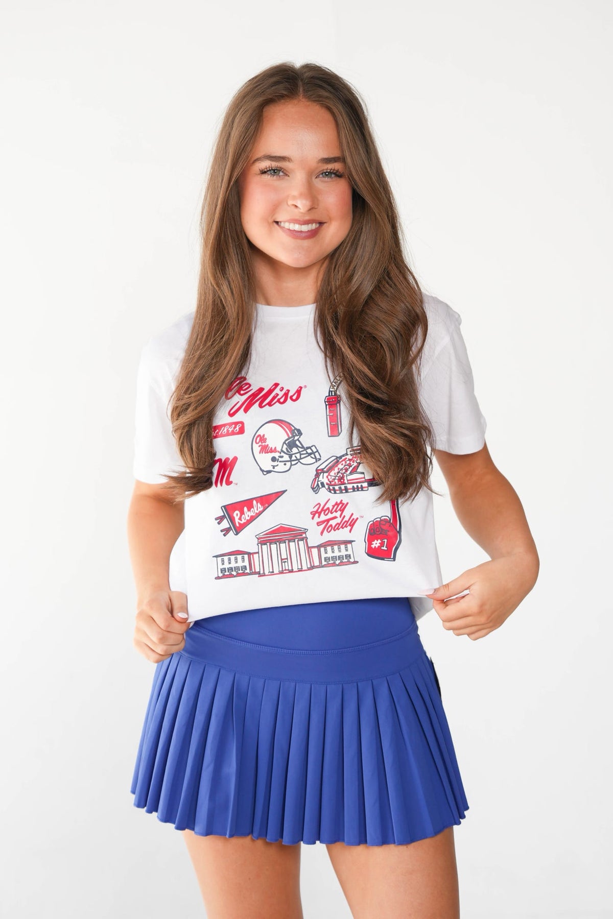A young girl with long brown hair stands against a white studio background wearing a white graphic T-shirt and a royal blue pleated mini skirt. The T-shirt features red and black collegiate-style graphics and text, including 'Miss' and 'Hwy.