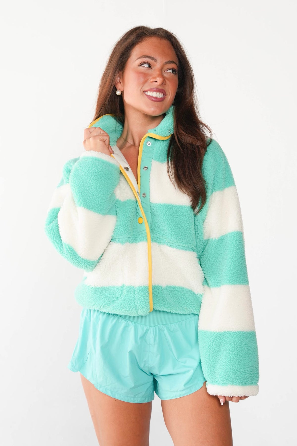 A woman modeling a matching fuzzy lounge set consisting of a turquoise and white striped quarter-zip fleece jacket with yellow trim and matching turquoise shorts, shot against a plain white studio background.
