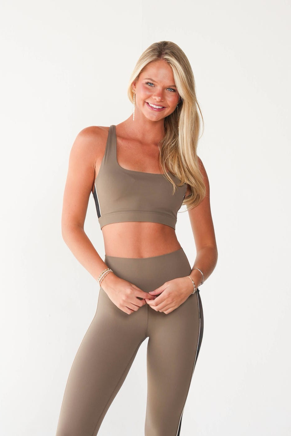 A woman modeling a matching taupe-brown activewear set consisting of a scoop-neck sports bra and high-waisted leggings with a black side stripe, paired with white sneakers against a plain white studio background.