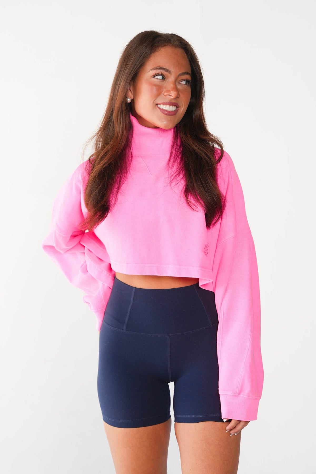 A woman modeling a bright pink long-sleeve crop top with a mock neck and navy blue high-waisted athletic shorts against a plain white background.