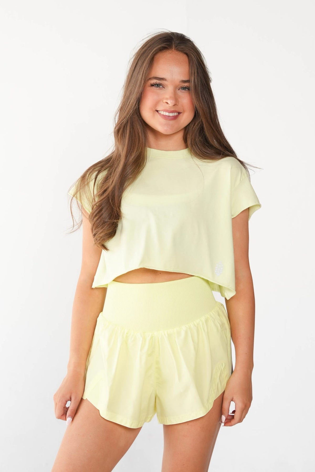 A young woman modeling a pale yellow two-piece lounge set consisting of a short-sleeve crop top and matching high-waisted shorts, set against a plain white studio background.