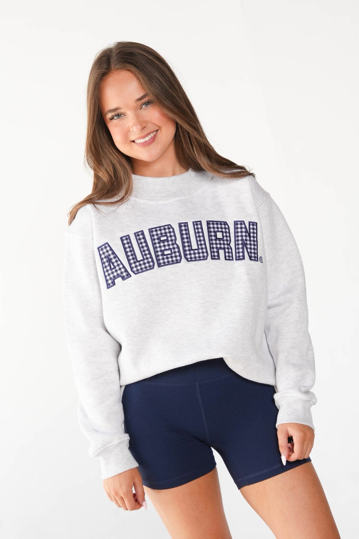 A young woman smiling and wearing a light gray heather crewneck sweatshirt with 'AUBURN' printed across the chest in navy blue gingham lettering, paired with navy blue athletic shorts, against a plain white studio background.