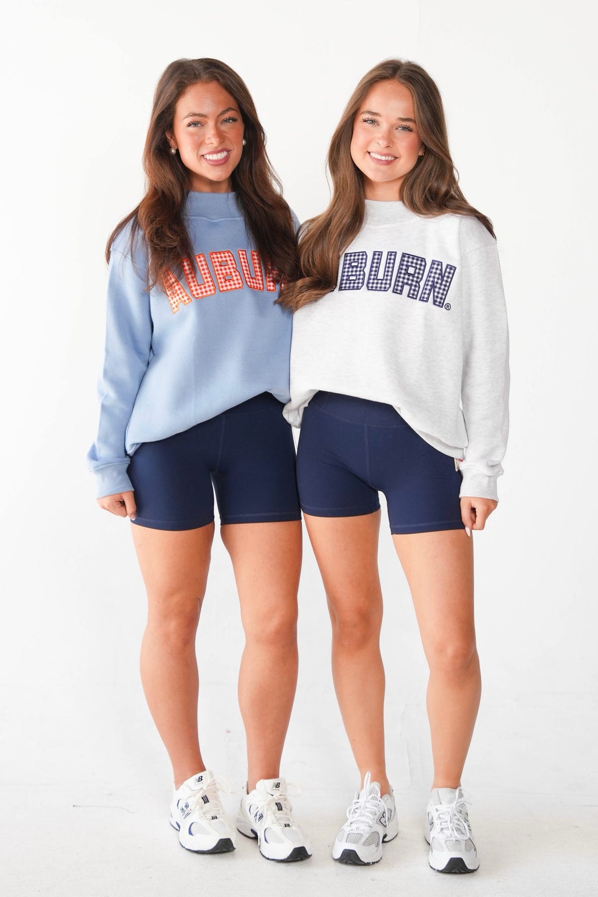 Two young women standing side-by-side in a studio setting, wearing Auburn athletic sweatshirts and navy blue bike shorts. The woman on the left wears a light blue sweatshirt with orange lettering, and the woman on the right wears a white sweatshirt with navy lettering.
