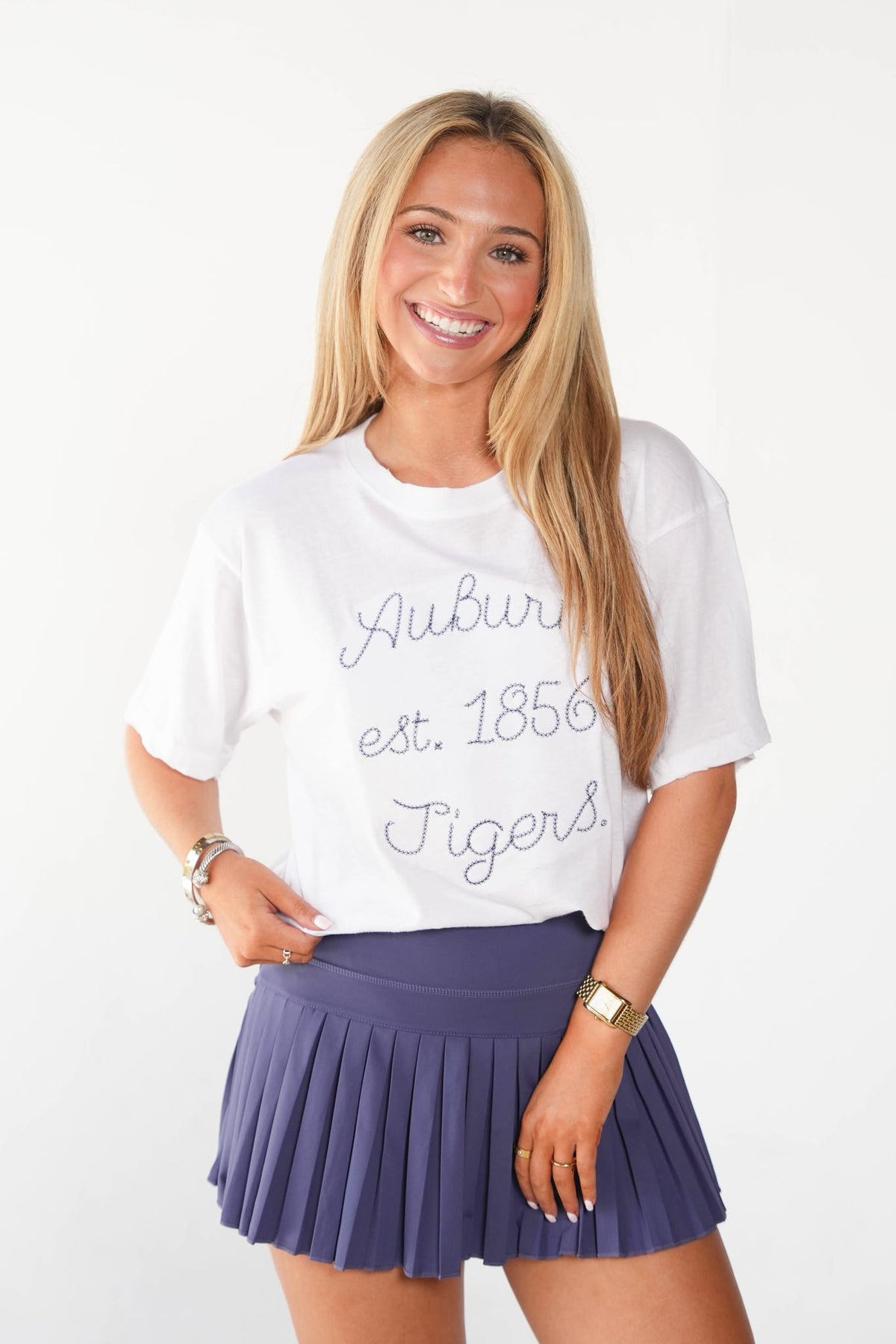 A smiling blonde woman wearing a white short-sleeve graphic T-shirt with 'Auburn est. 1856 Tigers' in gray script and a pleated purple tennis skirt, posed against a clean white studio background.