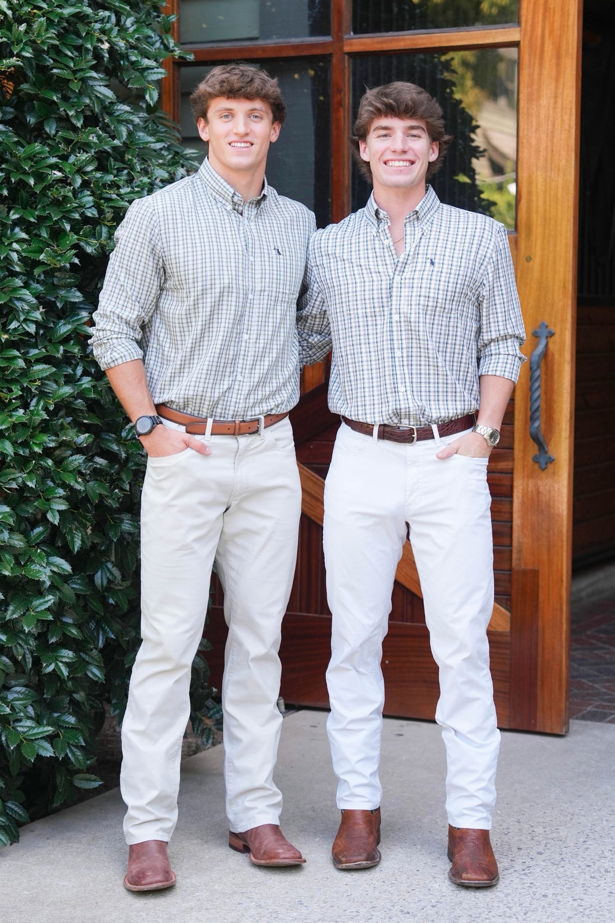Two young men standing outdoors in front of a wooden door and green hedge, wearing matching light-colored plaid button-down shirts, beige pants, and brown boots.