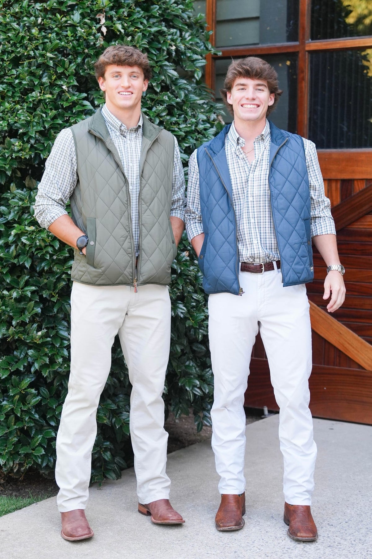 Two men standing outdoors in front of a green hedge and wooden door, each wearing a quilted vest over a plaid button-up shirt and white pants.