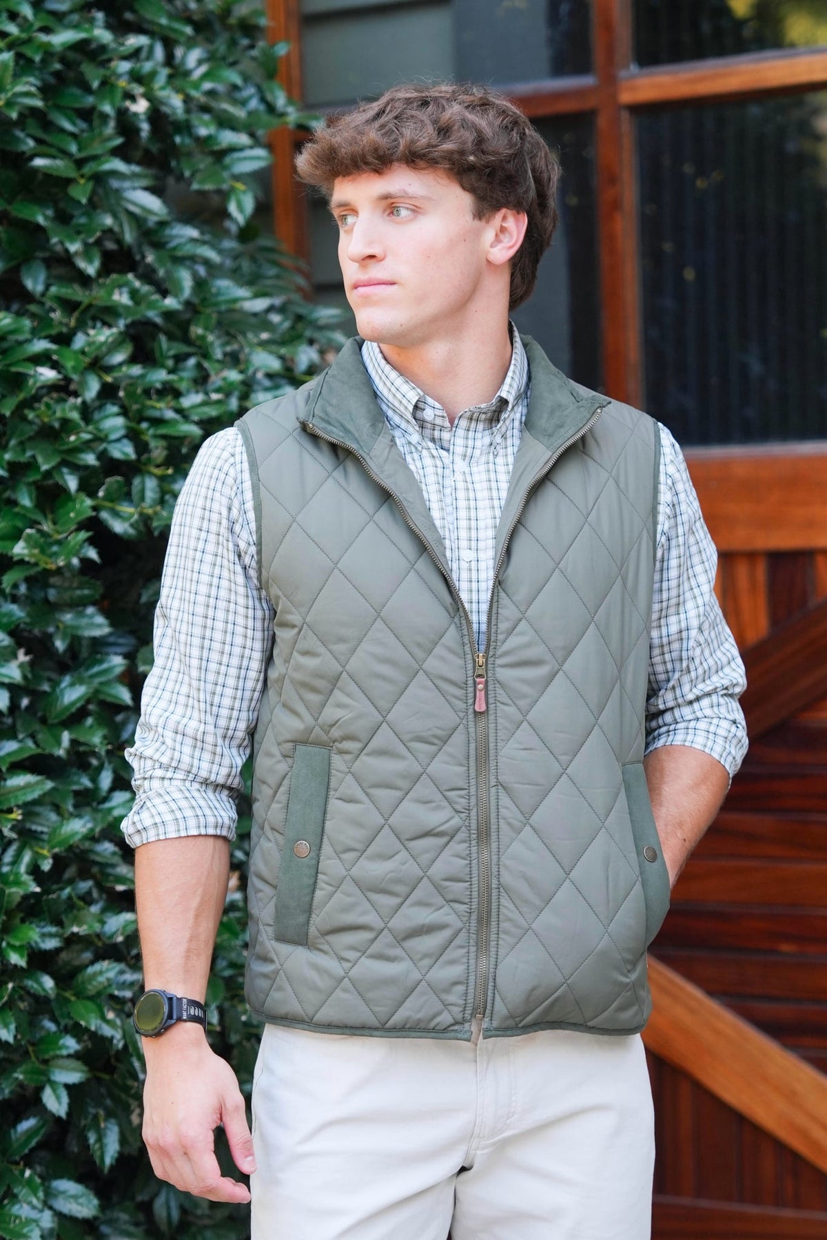 A man wearing an olive green diamond-quilted zip-up vest over a light blue and white checkered shirt, paired with beige pants, standing outdoors in front of a wooden door and green foliage.