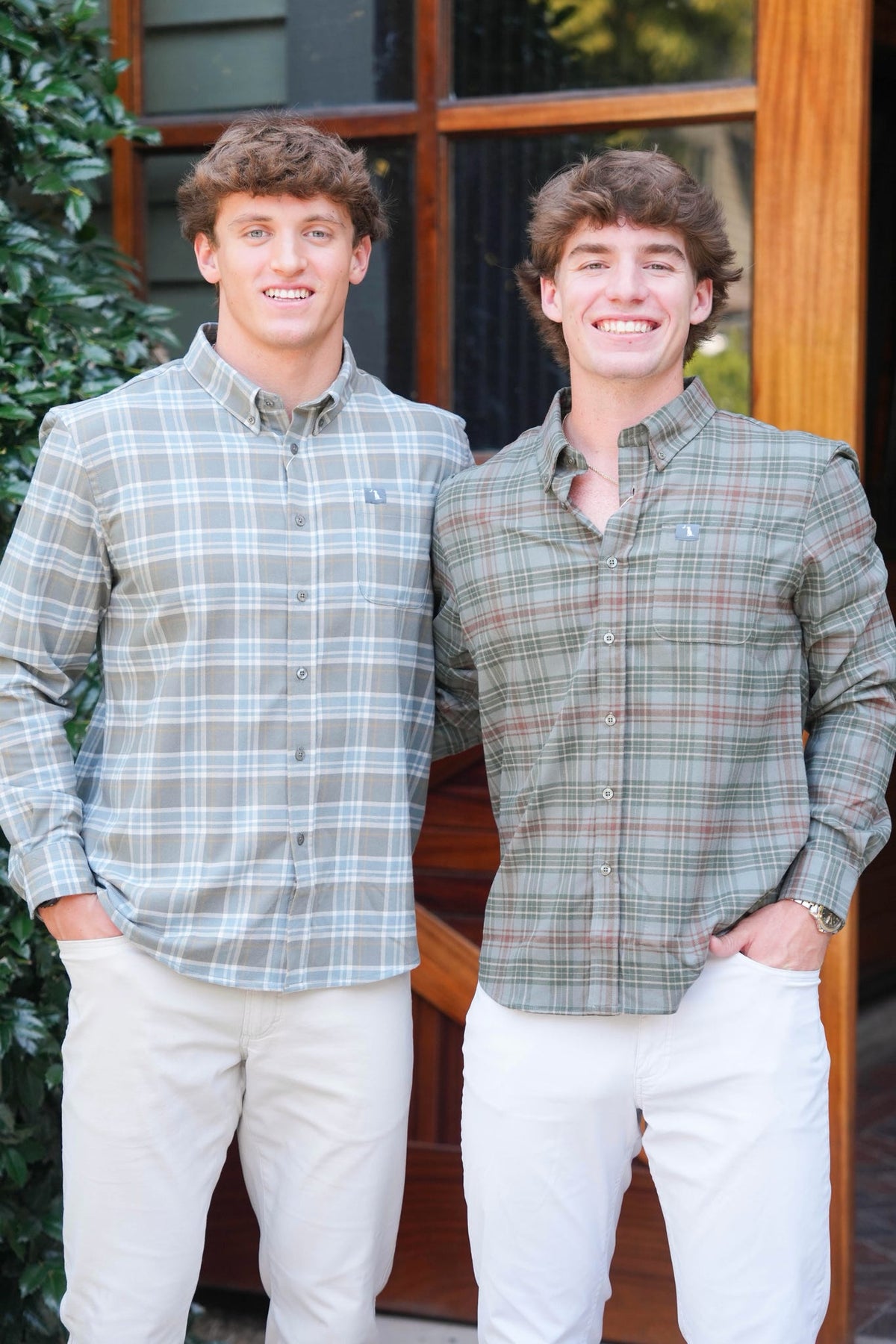 Two young men smiling and standing side-by-side outdoors in front of a wooden door. The man on the left wears a light gray and white plaid button-down shirt with beige pants, and the man on the right wears a muted green and brown plaid button-down shirt with white pants.