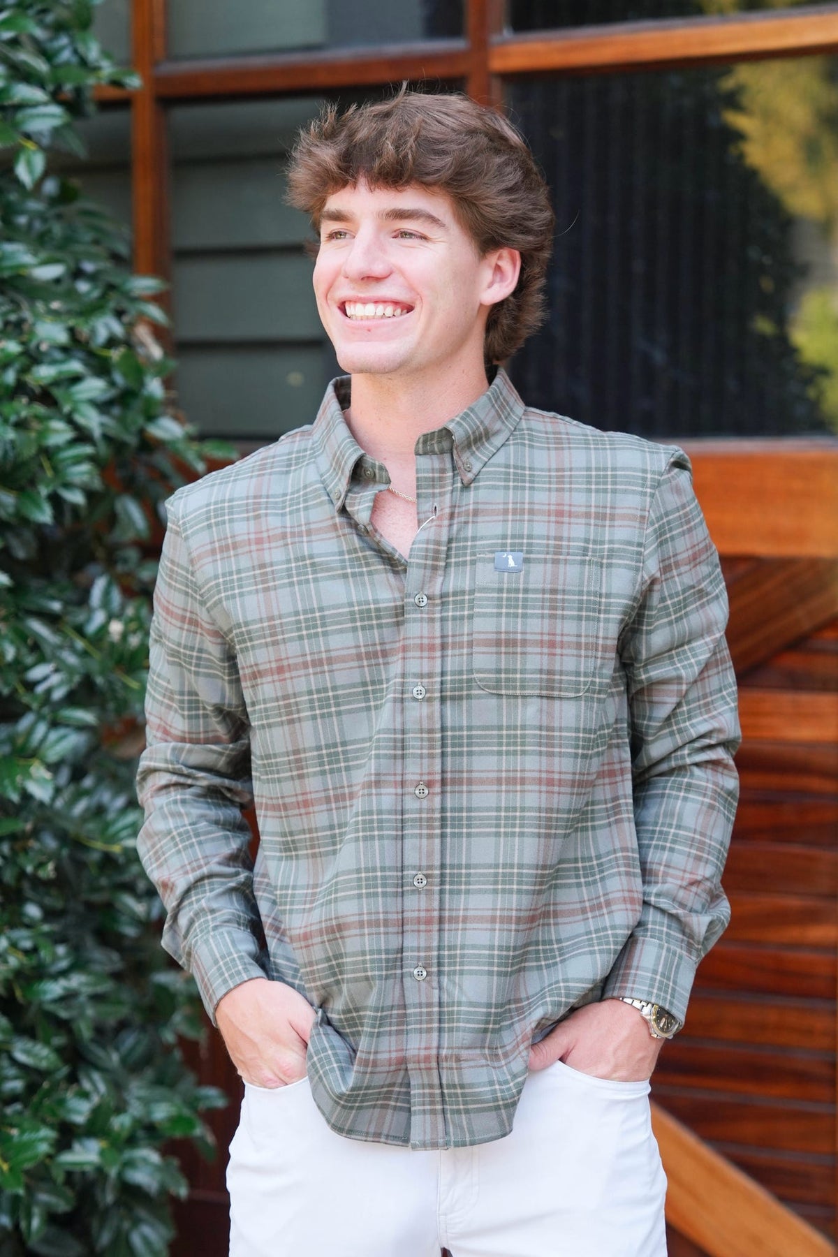 A smiling young man wearing a long-sleeve plaid button-down shirt in muted green, beige, and brown tones, paired with white pants, standing outdoors in front of a wooden building.