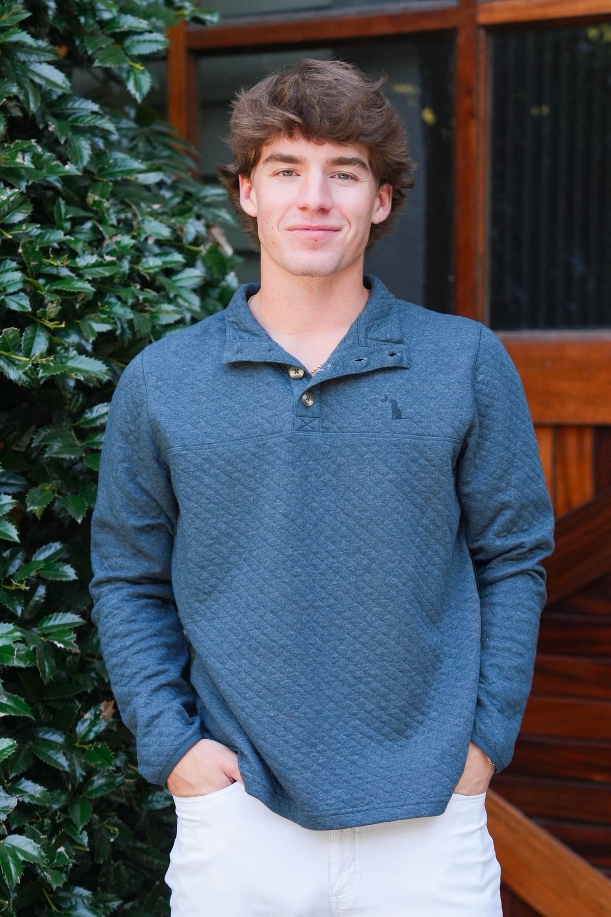 A young man with wavy brown hair stands outdoors wearing a textured slate blue long-sleeve polo shirt and white pants, with green foliage and a wooden door in the background.