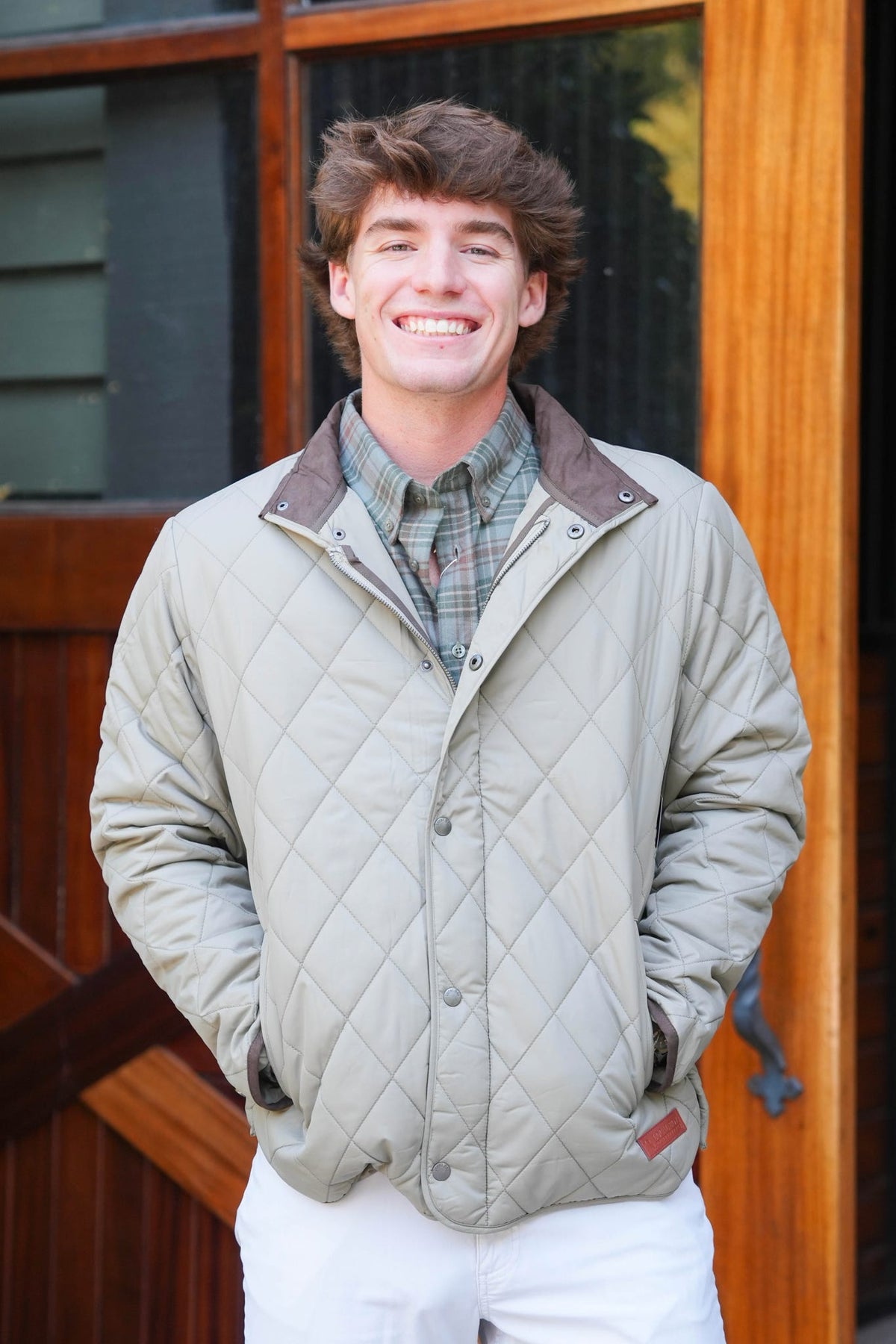 A smiling young man wearing a light beige diamond-quilted jacket over a green plaid shirt and white pants, standing in front of a rustic wooden door.