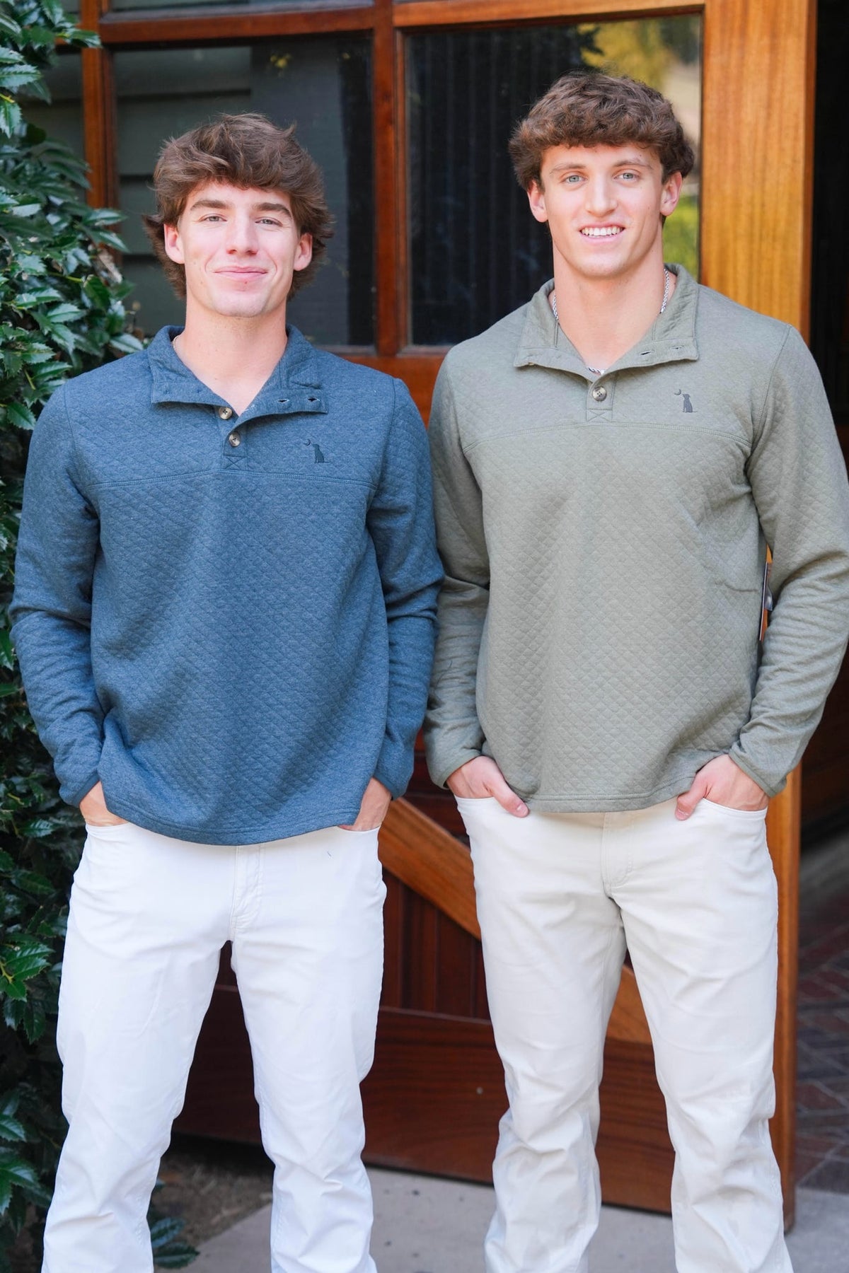 Two young men standing side-by-side wearing matching textured quarter-zip pullovers from Polo Ralph Lauren, one in slate blue and the other in sage green, paired with white pants, posed in front of a wooden door.