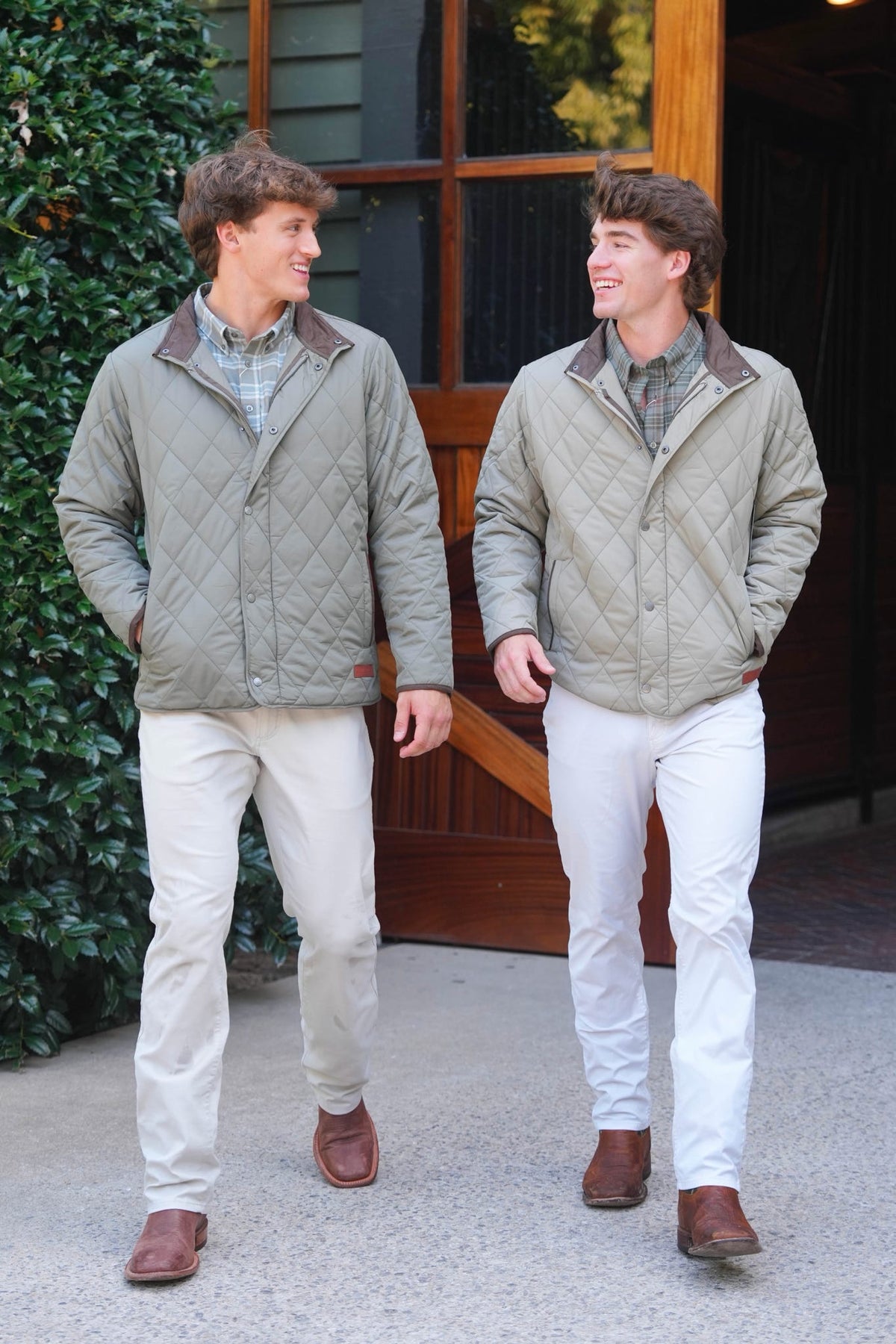 Two men walking outdoors wearing matching quilted light olive green jackets with brown collar trim, paired with light pants and brown shoes, in front of a rustic wooden building.