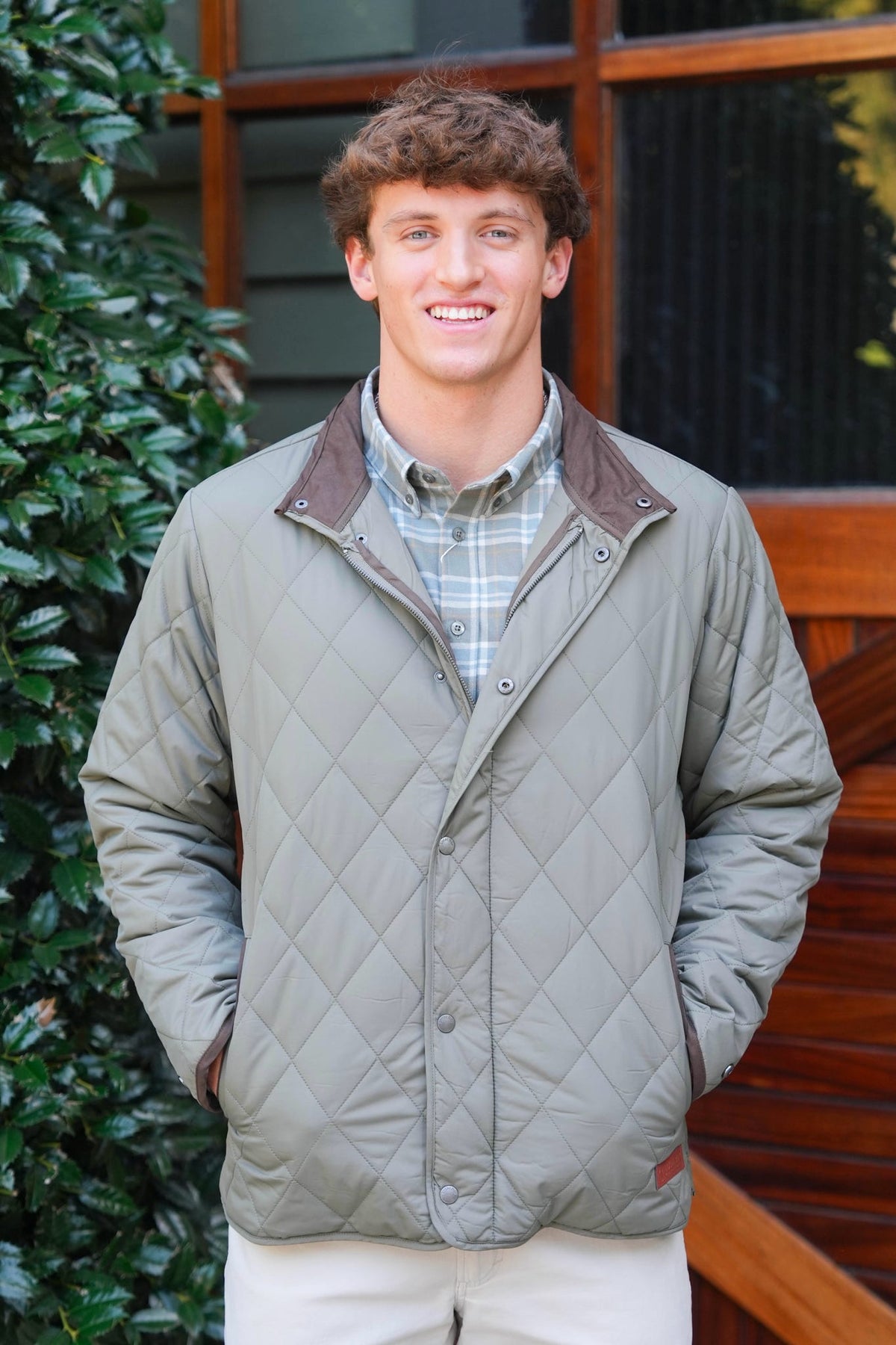 A smiling young man wearing a sage green diamond-quilted jacket with brown collar and pocket trim, layered over a plaid button-down shirt and cream pants, standing outdoors in front of a wooden door and green foliage.