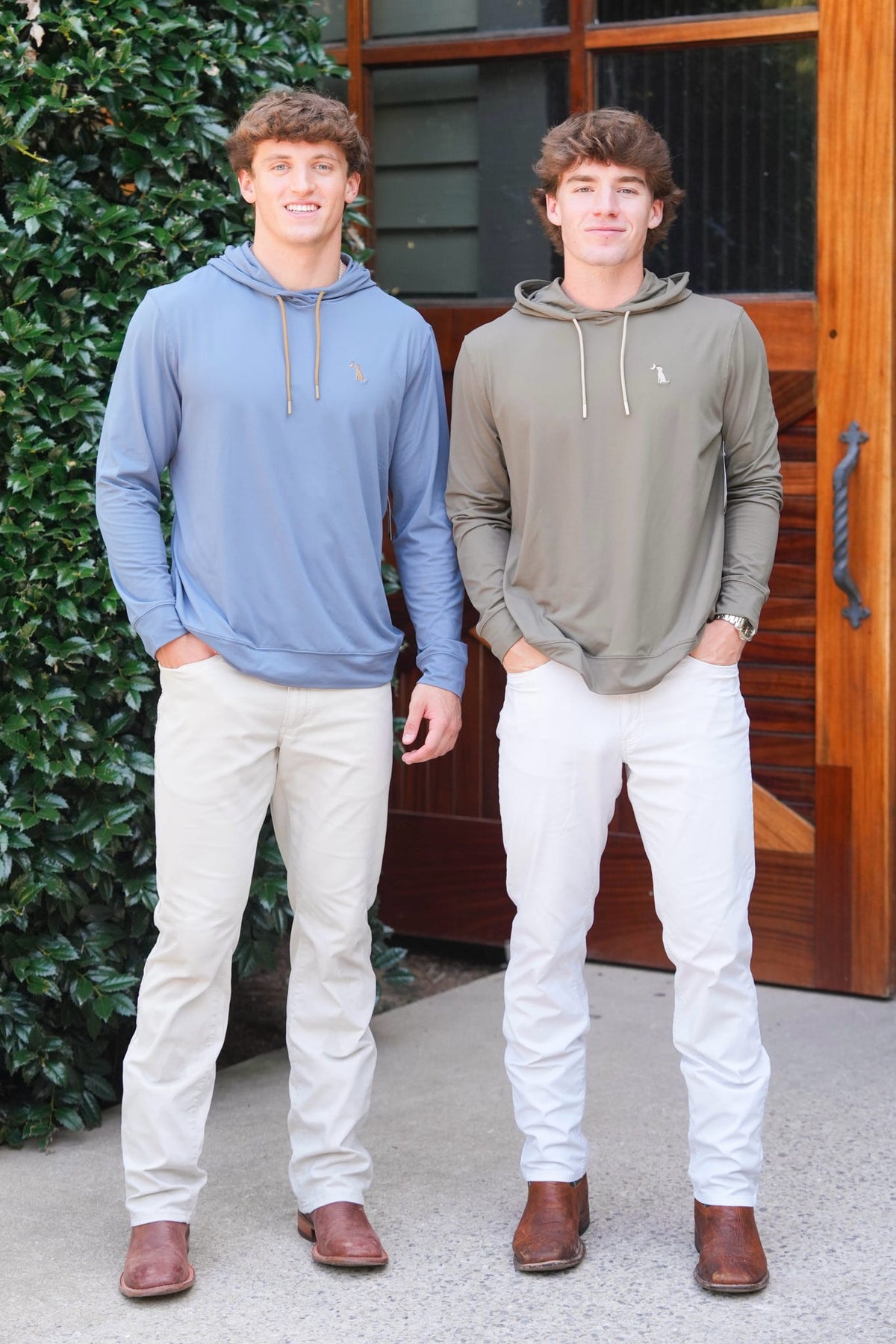 Two young men standing outdoors in front of a wooden door and green hedge, wearing Polo Ralph Lauren hoodies. The man on the left wears a light blue hoodie with beige pants, and the man on the right wears an olive green hoodie with white pants.