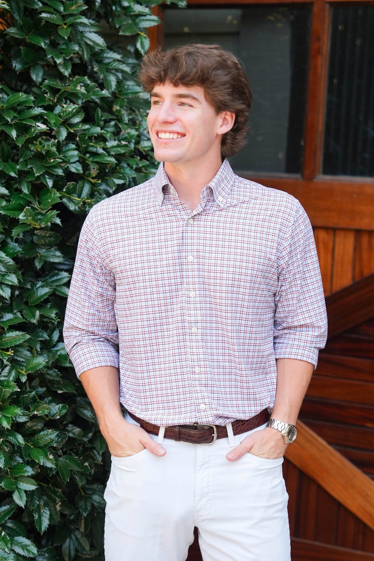 A smiling young man wearing a light pink and white checkered button-down shirt, white pants, and a brown belt, standing outdoors in front of a wooden door and green foliage.