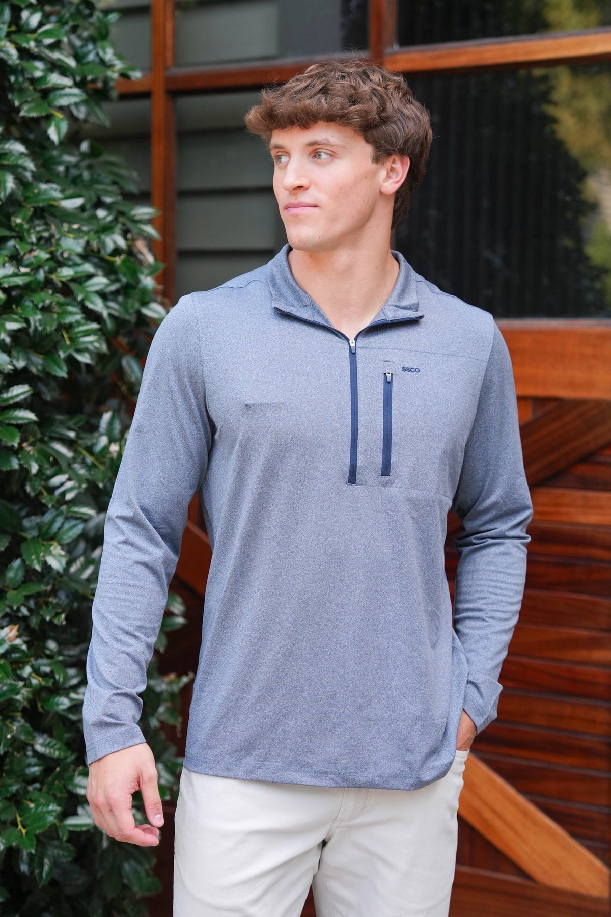 A man wearing a heathered slate blue long-sleeve quarter-zip pullover with a zippered chest pocket, paired with light beige pants, standing outdoors in front of a wooden building.