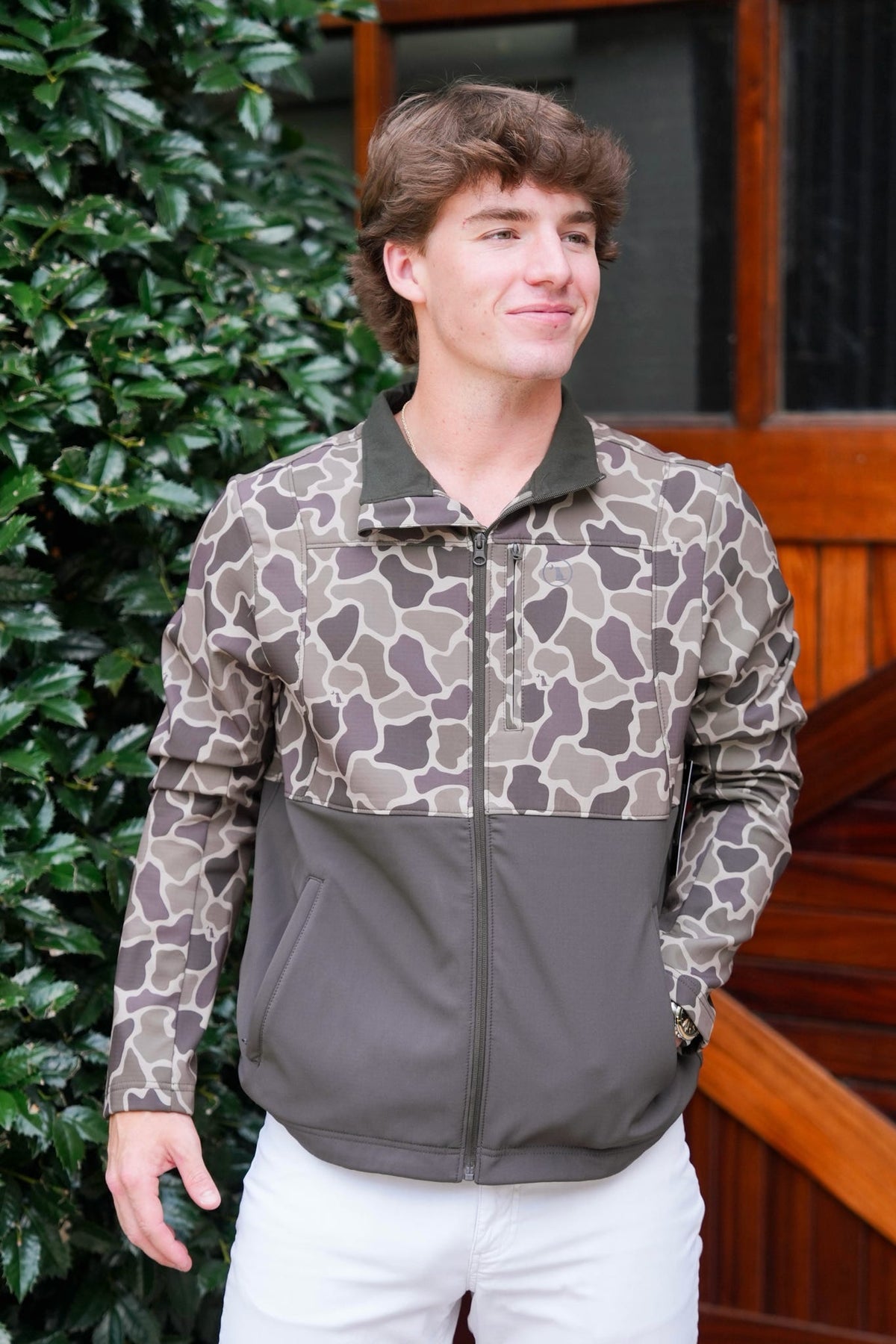 A man wearing a color-blocked zip-up jacket with a brown and beige animal print upper section and a solid dark brown lower section, paired with white pants, standing outdoors in front of a wooden door and green foliage.