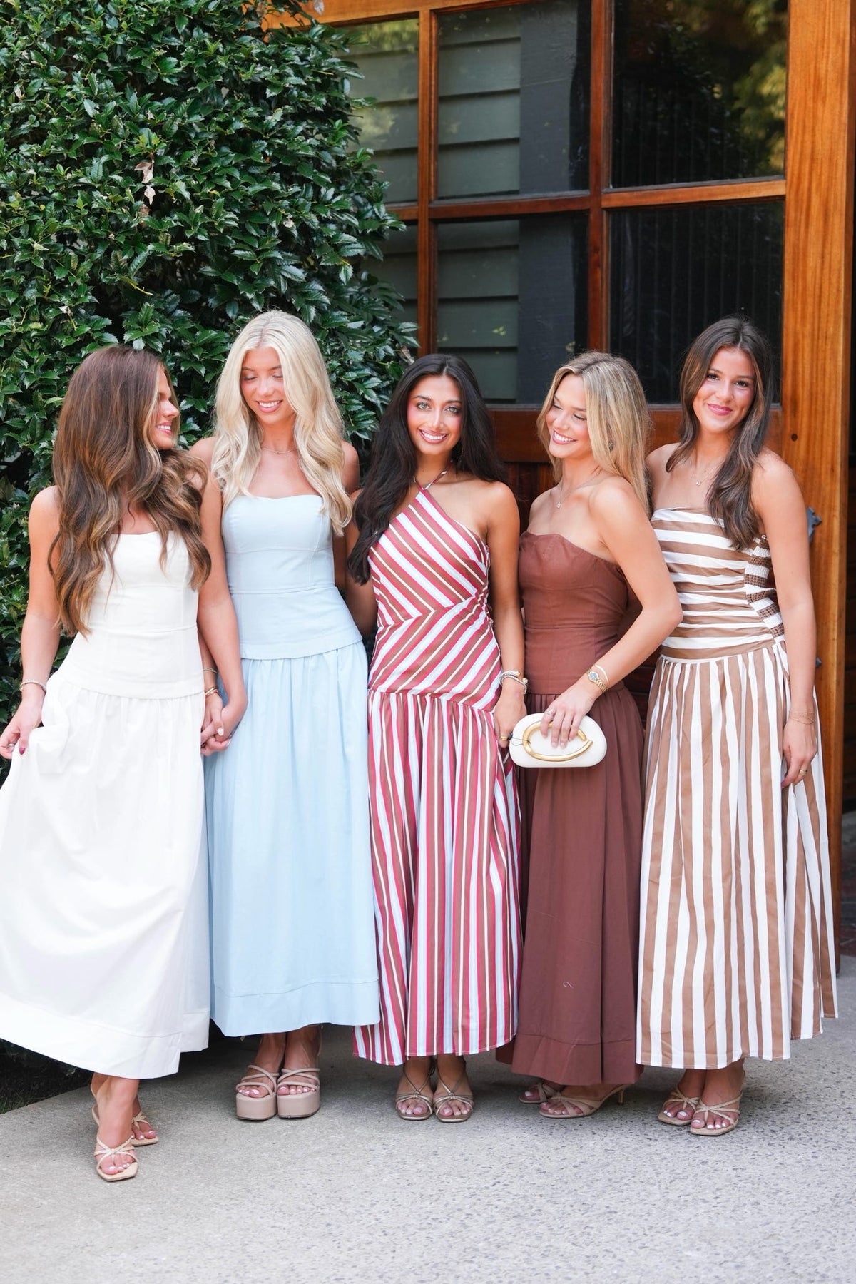 Five women standing together in front of a wooden door and greenery, wearing coordinated sleeveless dresses in white, light blue, red-and-white stripes, brown, and beige-and-white stripes.