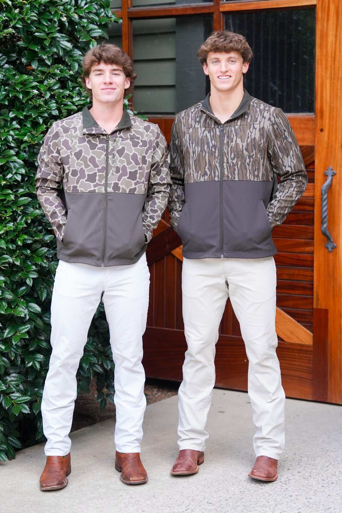 Two young men standing outdoors in front of a wooden door and greenery, wearing matching zip-front jackets with a brown and gray abstract pattern on the upper half and solid dark gray on the lower half, paired with white pants and brown shoes.