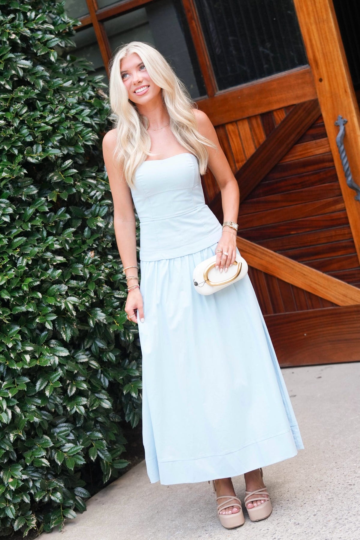 A woman with long blonde hair stands outdoors wearing a light blue sleeveless midi dress, holding a small beige clutch purse. She is positioned in front of a green leafy hedge and a rustic wooden door.
