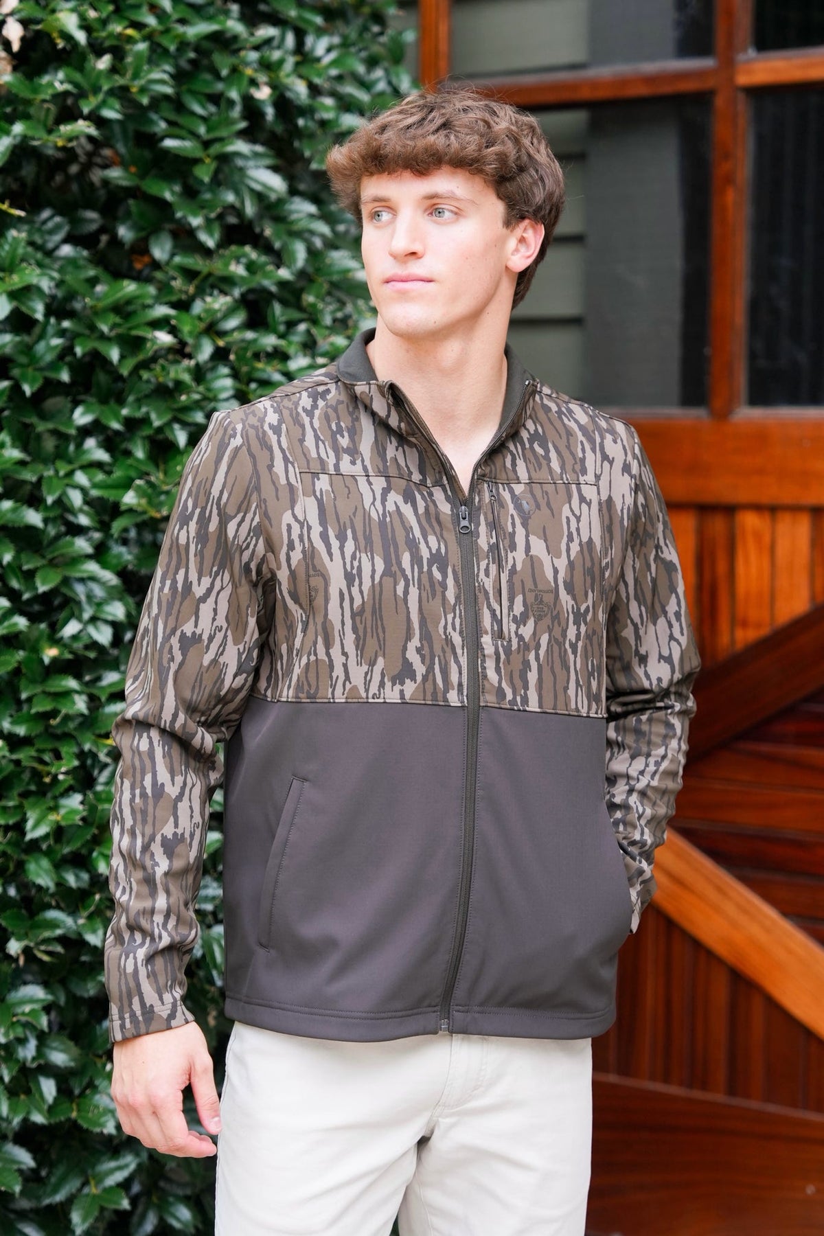 A man wearing a color-blocked zip-up jacket with a brown and beige camouflage pattern on the upper section and sleeves, and a solid dark gray lower section, standing outdoors in front of a wooden door and green hedge.